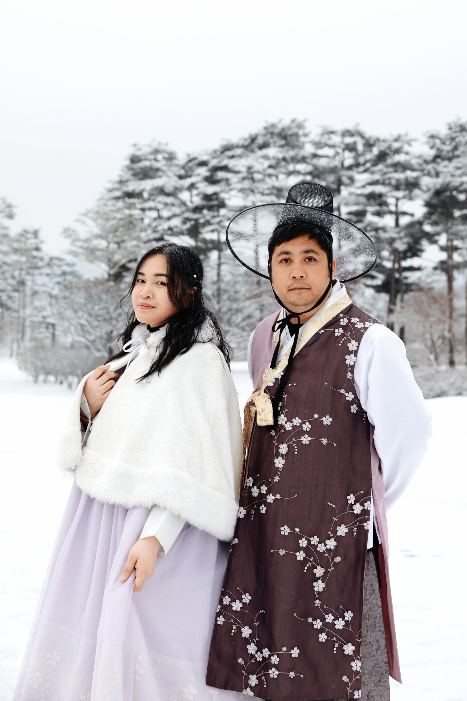 Two people in winter hanbok attire pose together in a snowy landscape with pine trees.