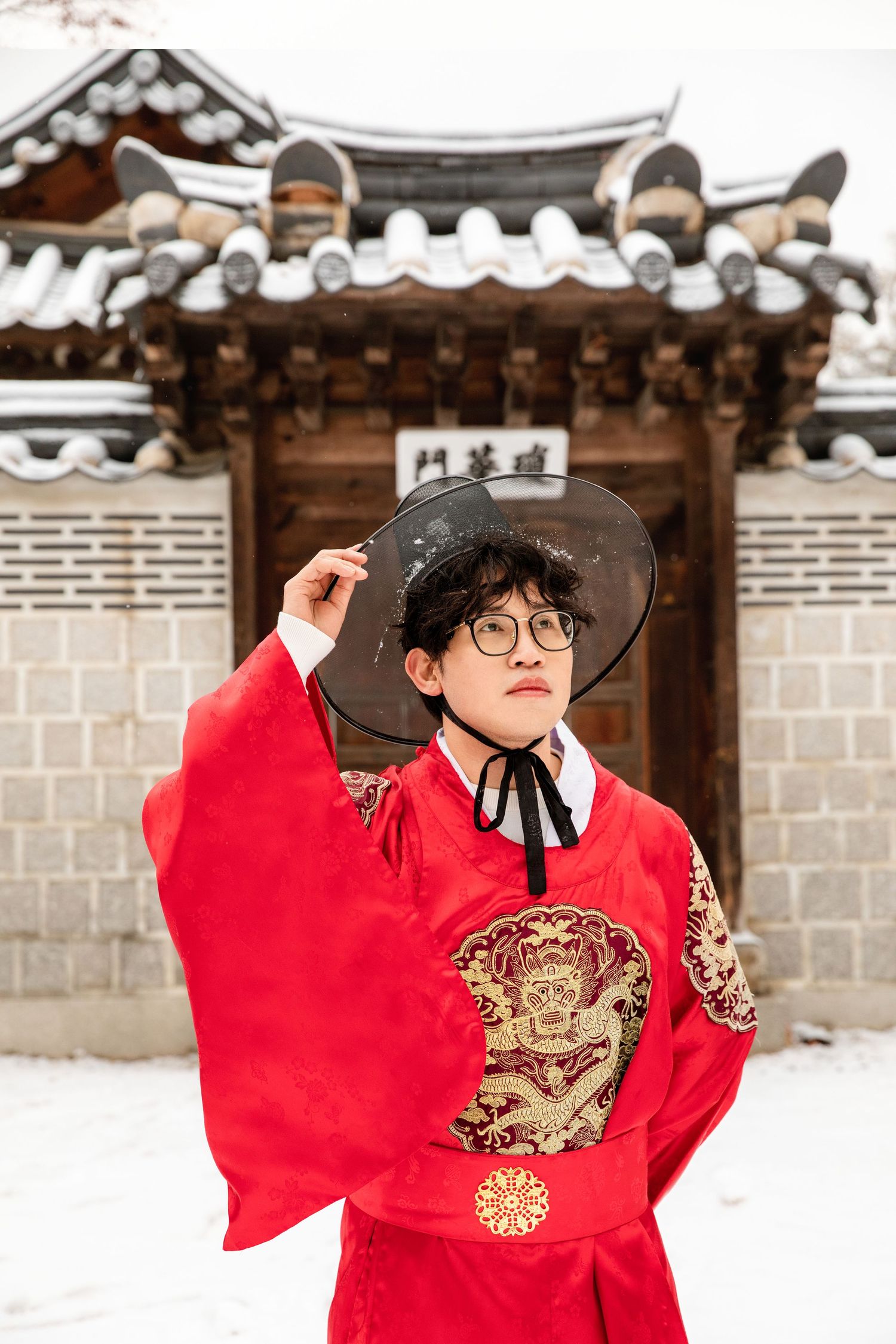Person wearing traditional red Korean hanbok and black hat poses in front of historic palace architecture in snowy setting.