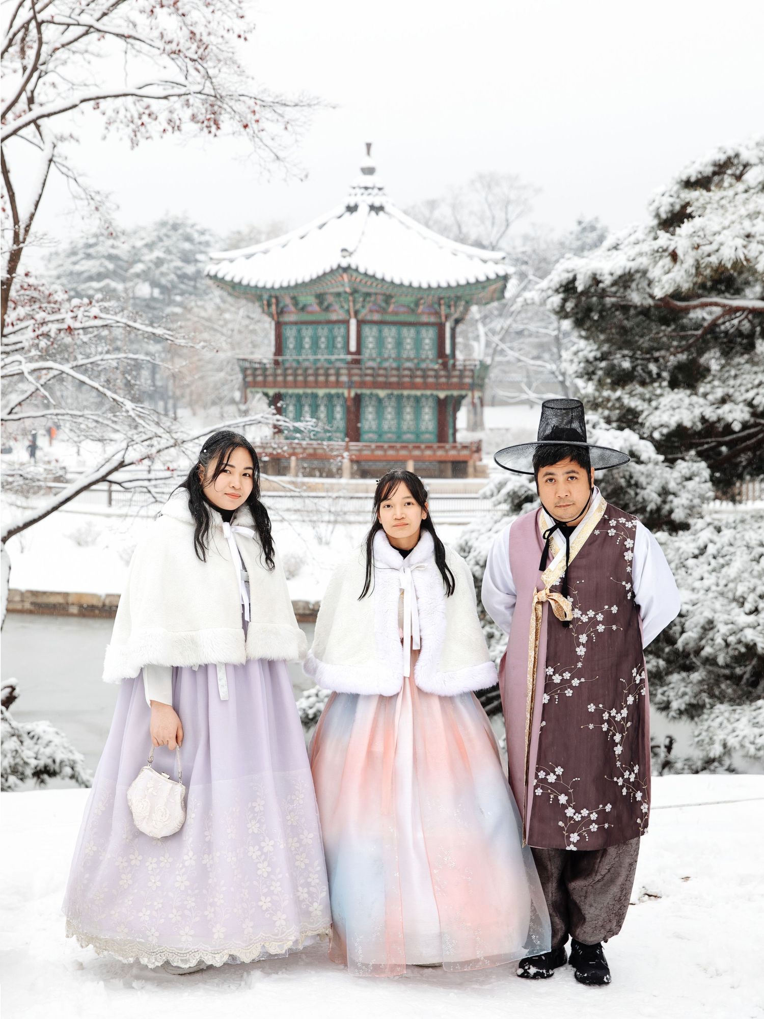 People wearing traditional Korean hanbok outfits pose in front of a snow-covered pagoda at a historic palace.