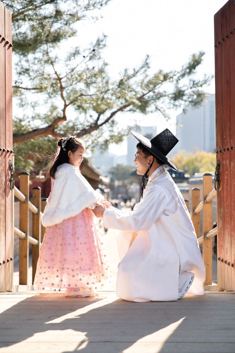 Two people in traditional Korean hanbok attire sitting together on a wooden bridge in sunlight.