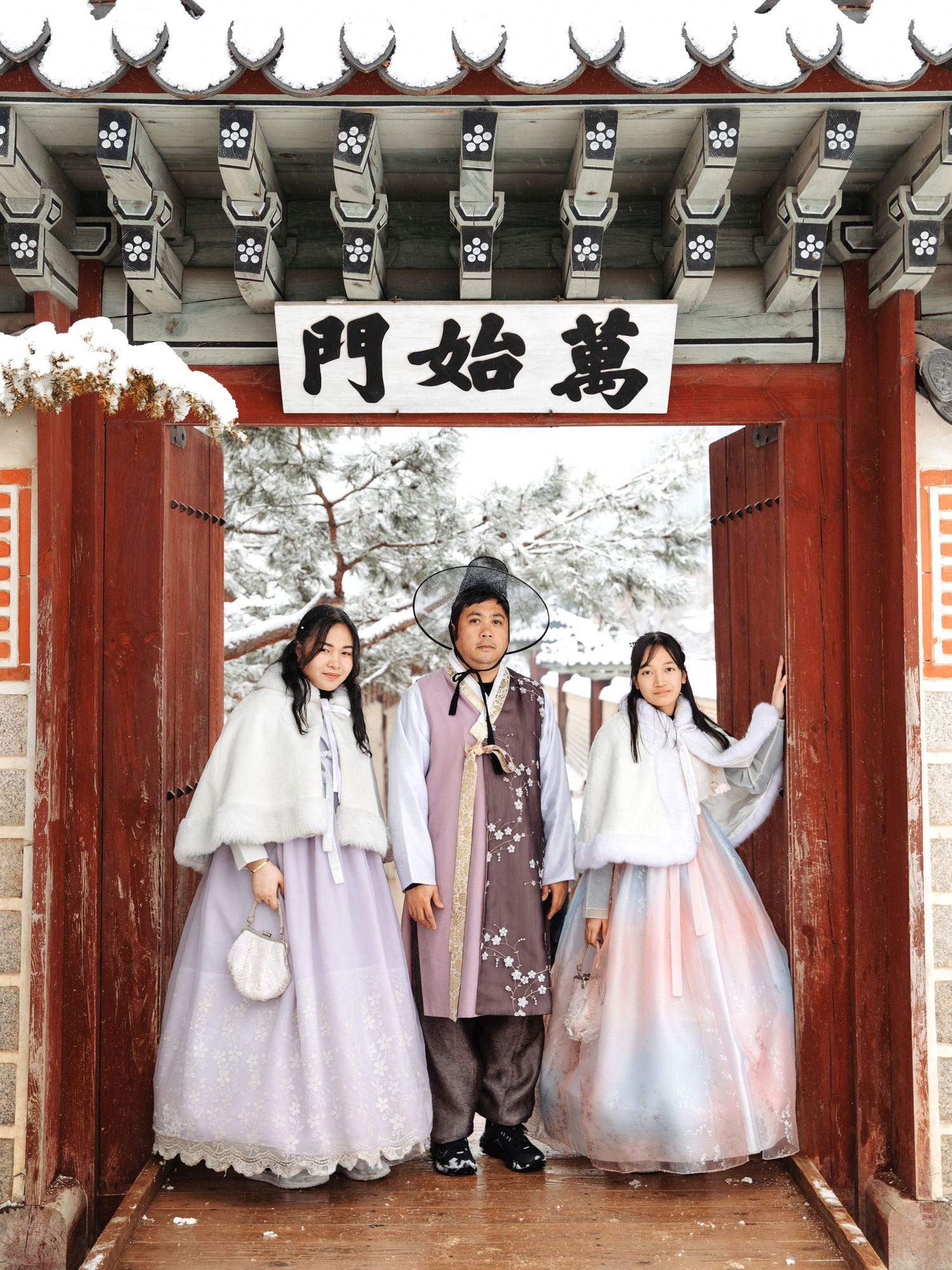 People in traditional Korean clothing pose under a red gateway entrance with snow-covered decorative roof.