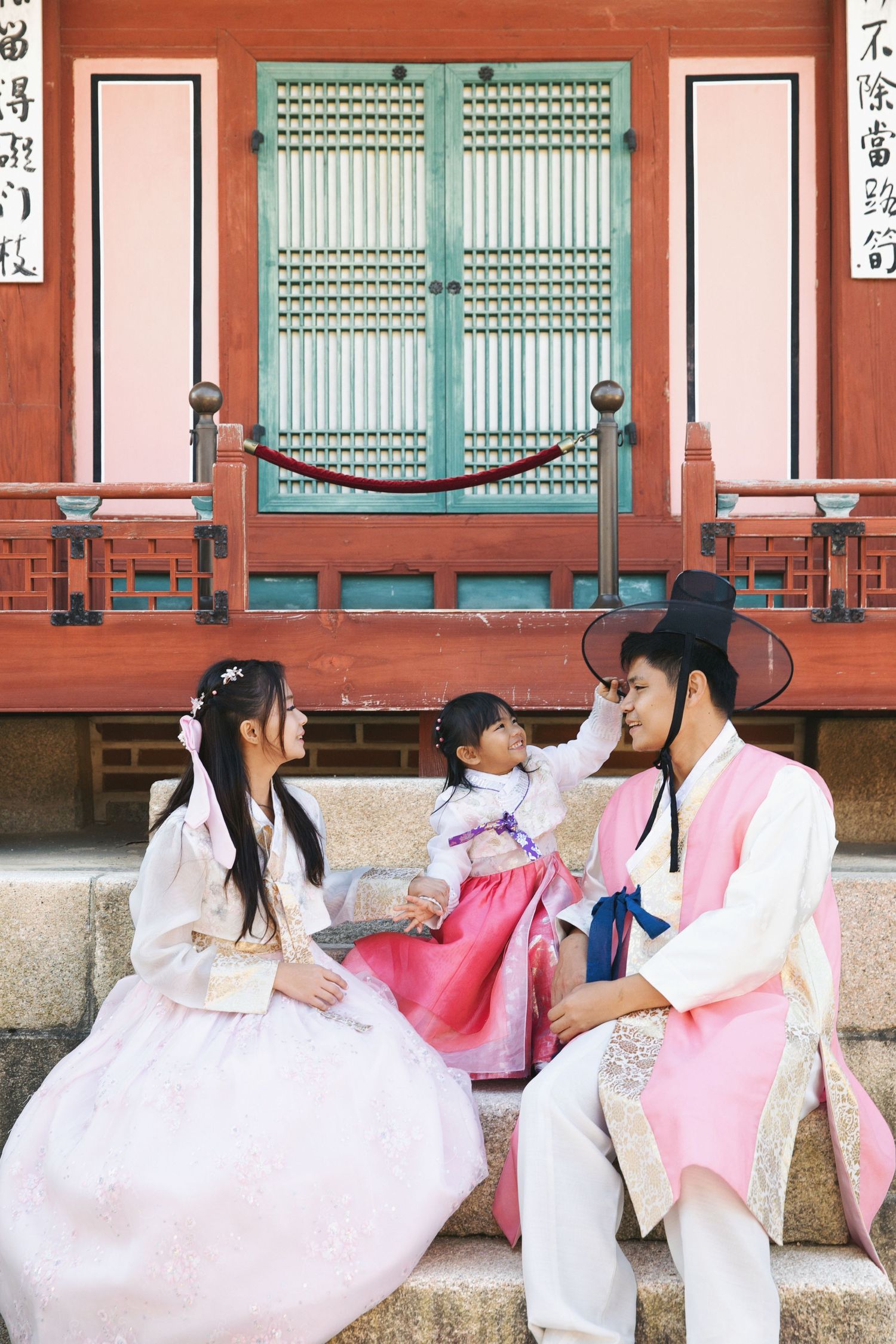 A family wearing traditional Korean hanbok outfits sits together on stone steps in front of a traditional building.