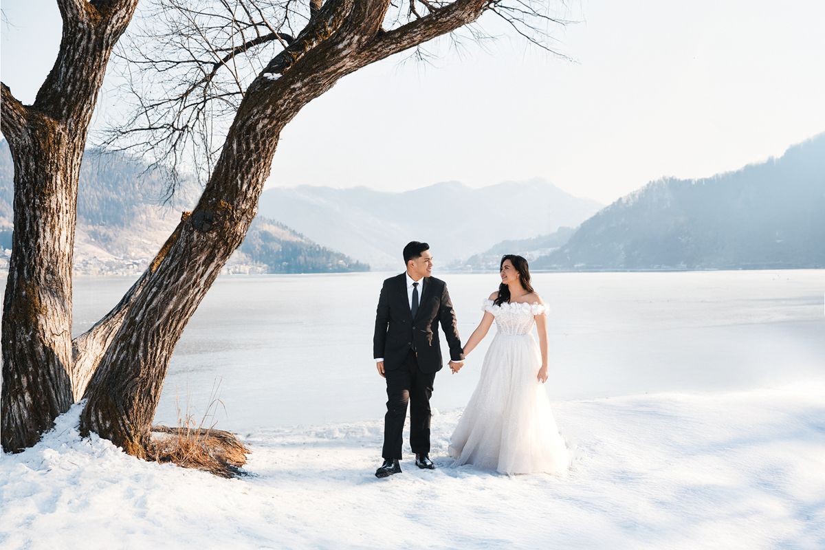A winter wedding couple stands holding hands beneath a bare tree by a snowy lakeside with mountains in the distance.