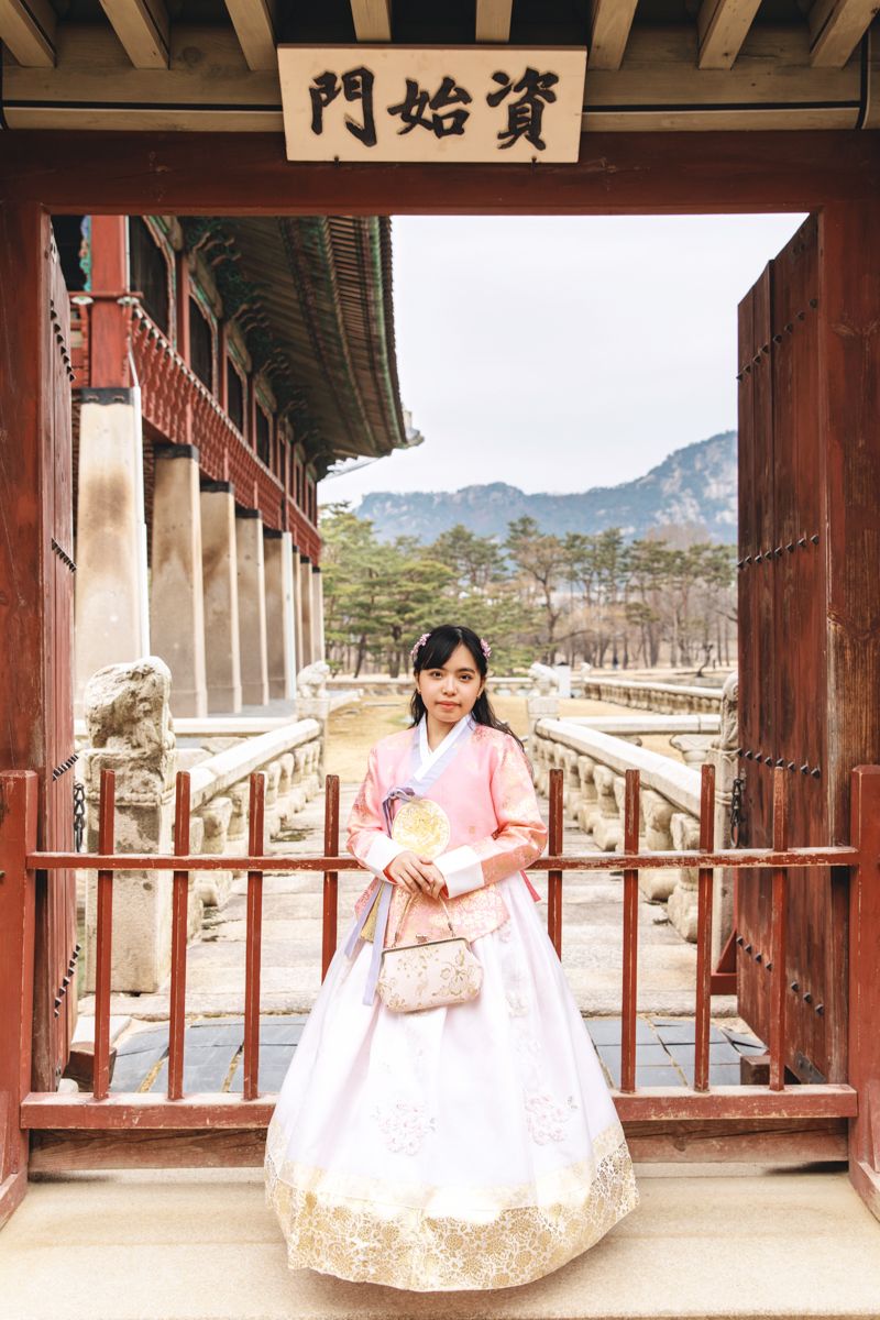 Traditional Korean hanbok-clad figure poses at ornate temple gateway with mountains and trees in background.