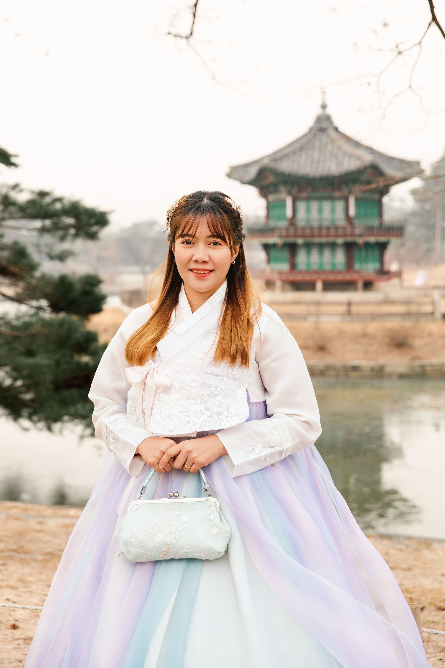 A person in a white and pastel hanbok dress poses by a lake with traditional Korean architecture.
