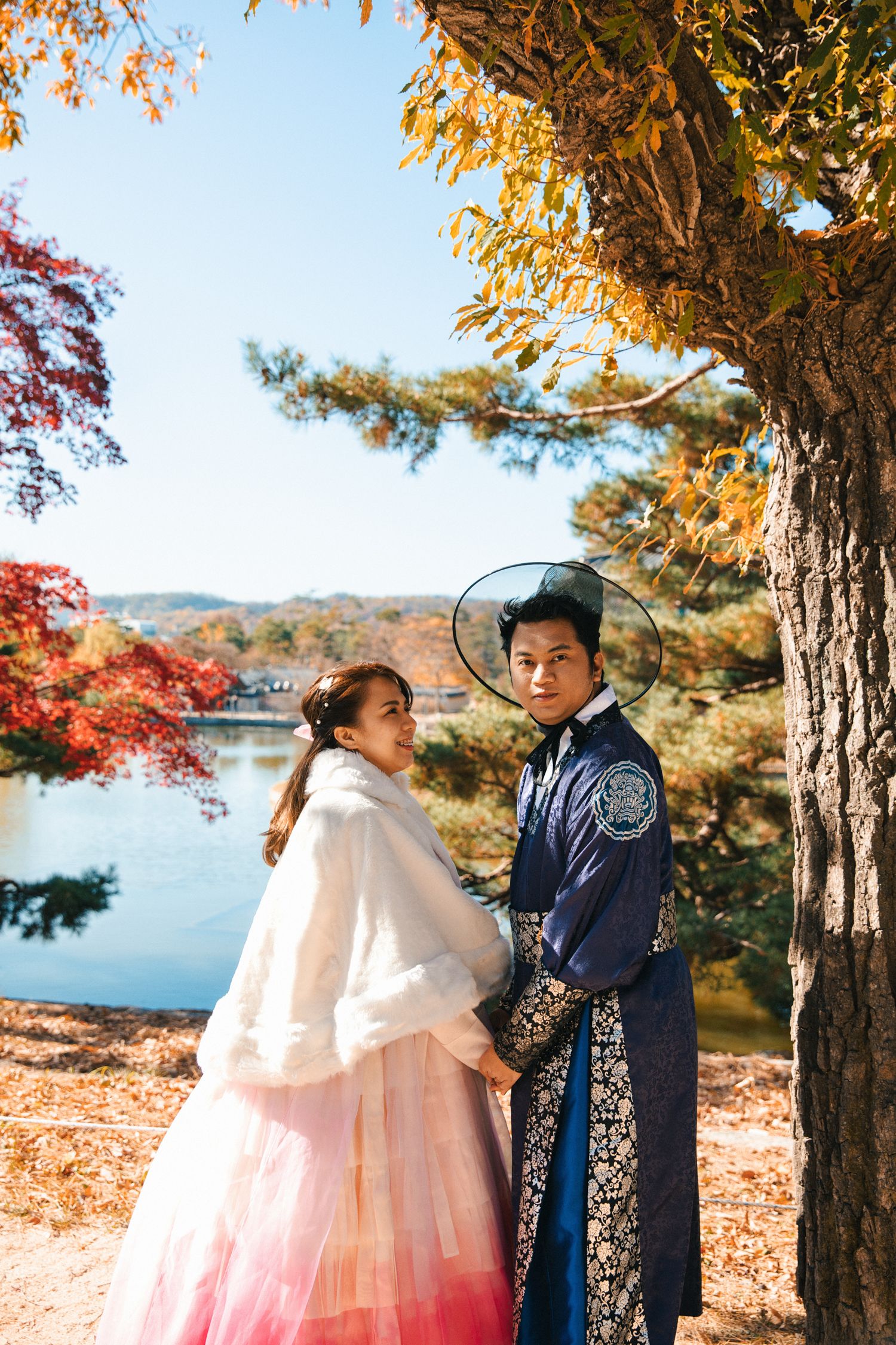 Couple in traditional Korean clothing poses by autumn trees with lake view in background.