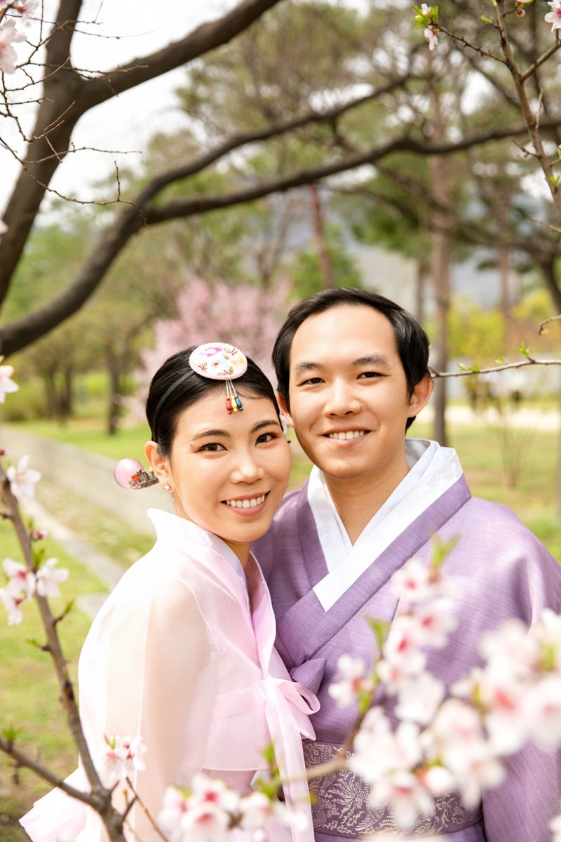 Two people in traditional Korean hanbok outfits embracing among spring cherry blossoms in a garden setting.