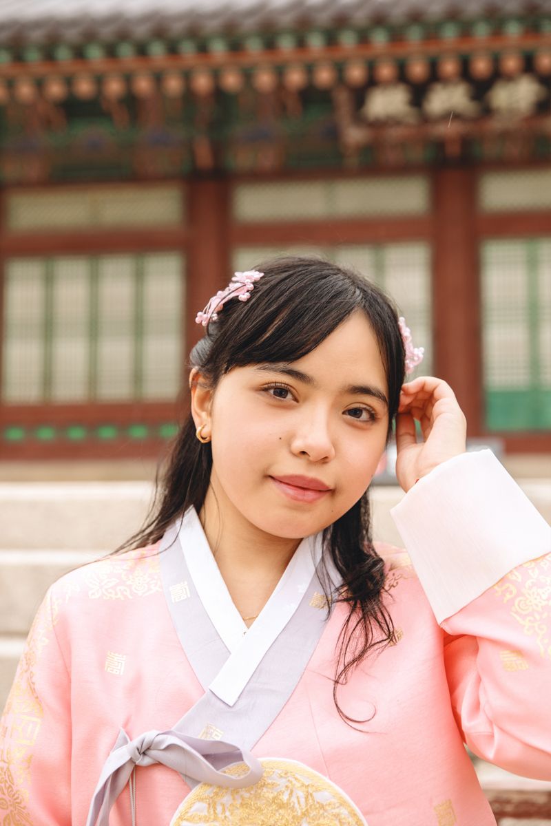Traditional Korean hanbok outfit in soft pink color with white collar worn at a temple with ornate architecture visible.