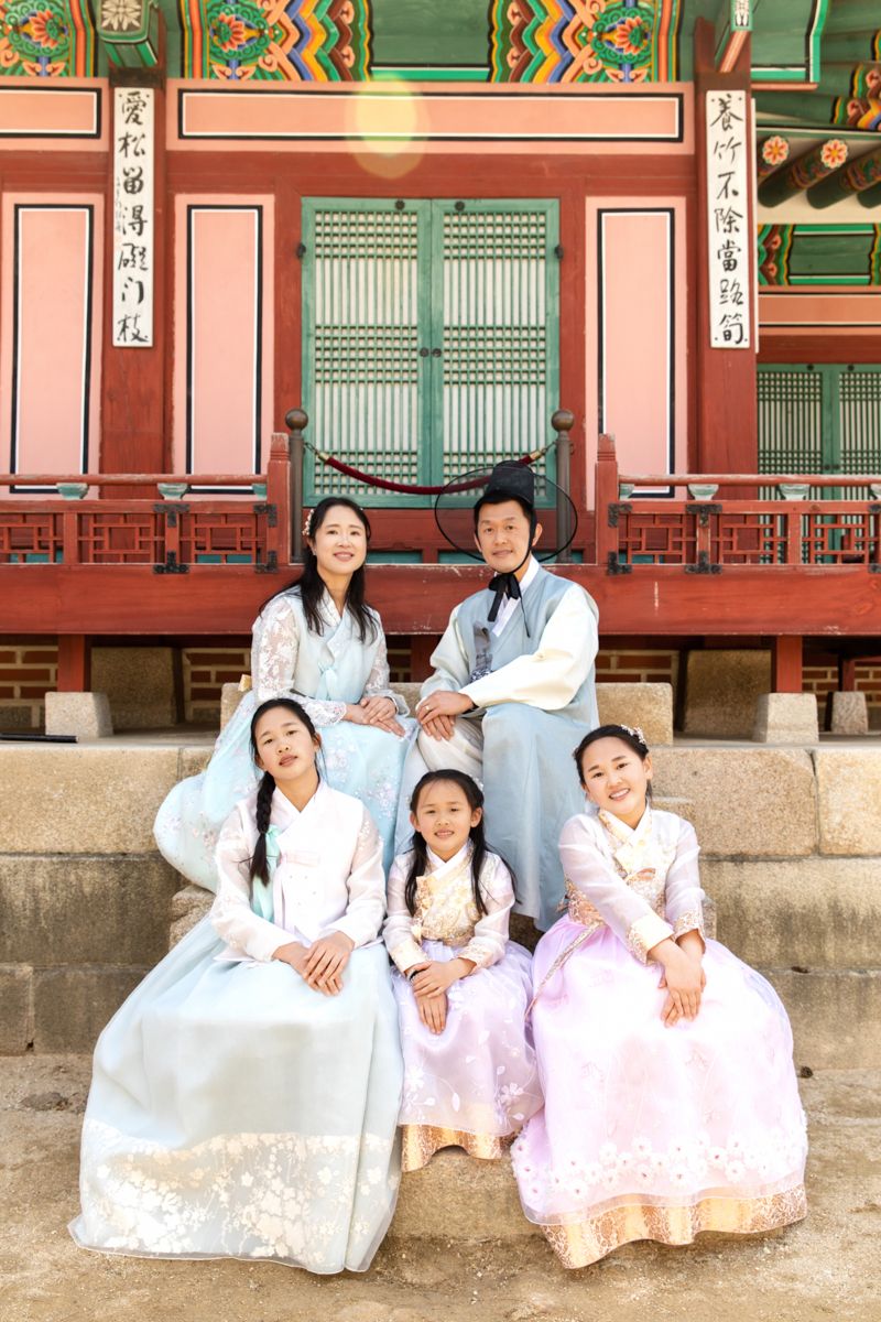 Group wearing traditional Korean hanbok attire seated on stone steps at ornate temple with colorful painted details.