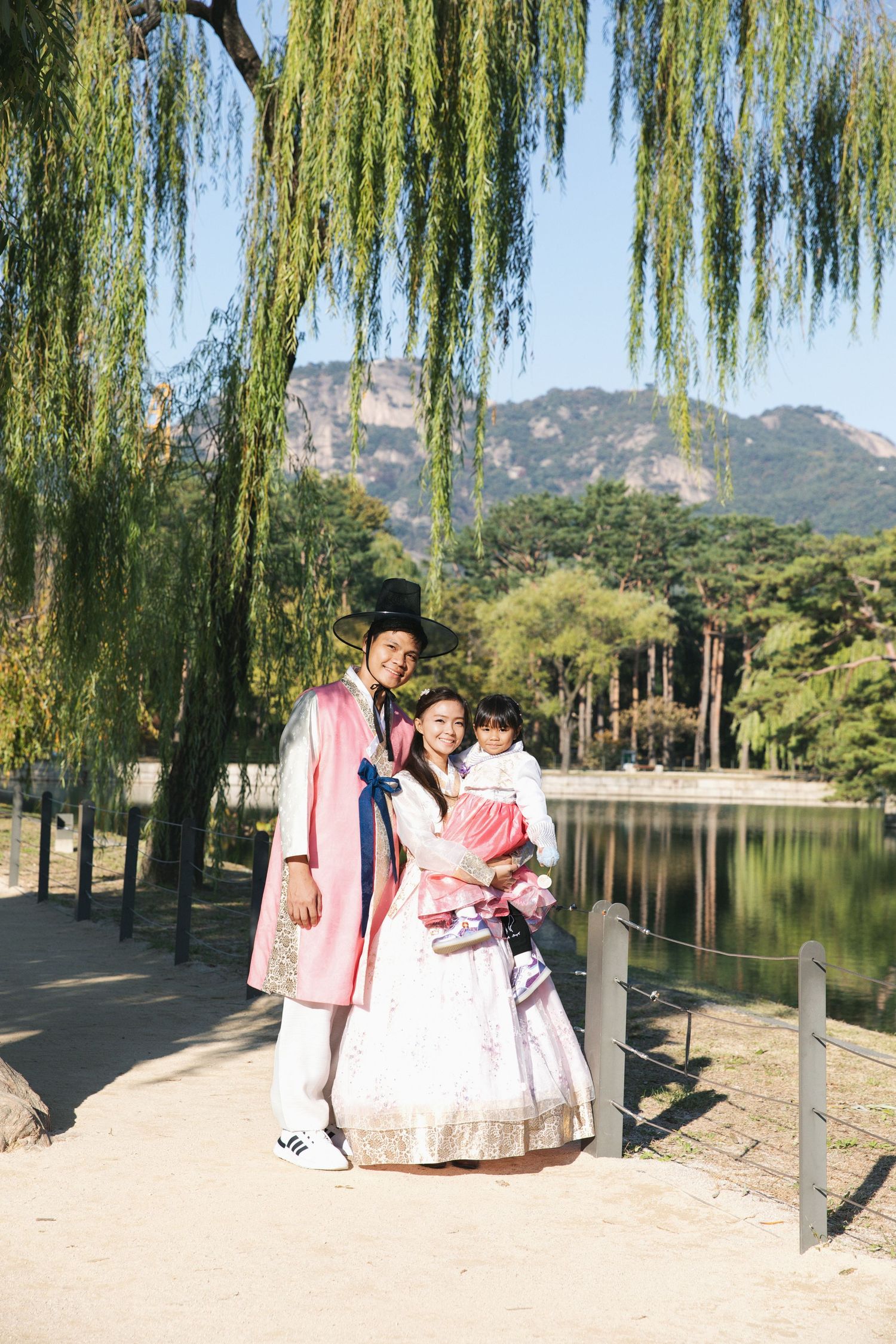 Family in traditional Korean hanbok attire posing beneath weeping willow trees by lakeside.