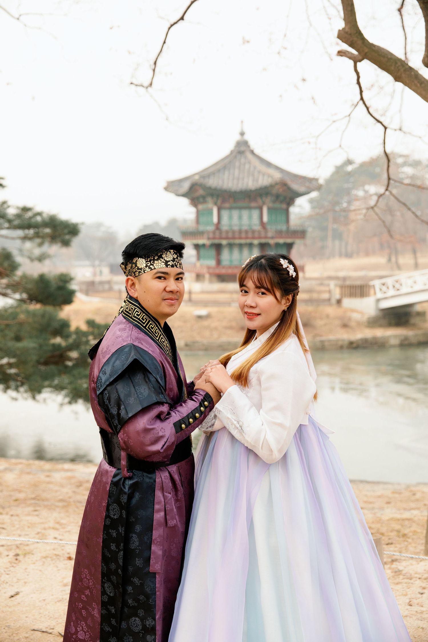 A couple in contrasting traditional Korean outfits poses by a lakeside pavilion.