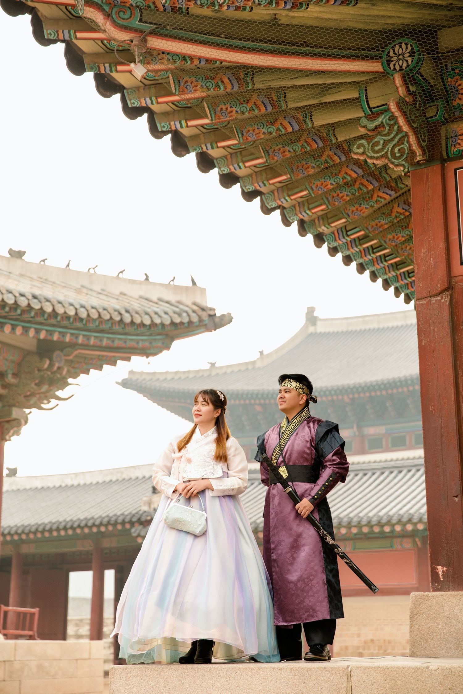 A couple in traditional Korean outfits poses together under ornate palace architecture.