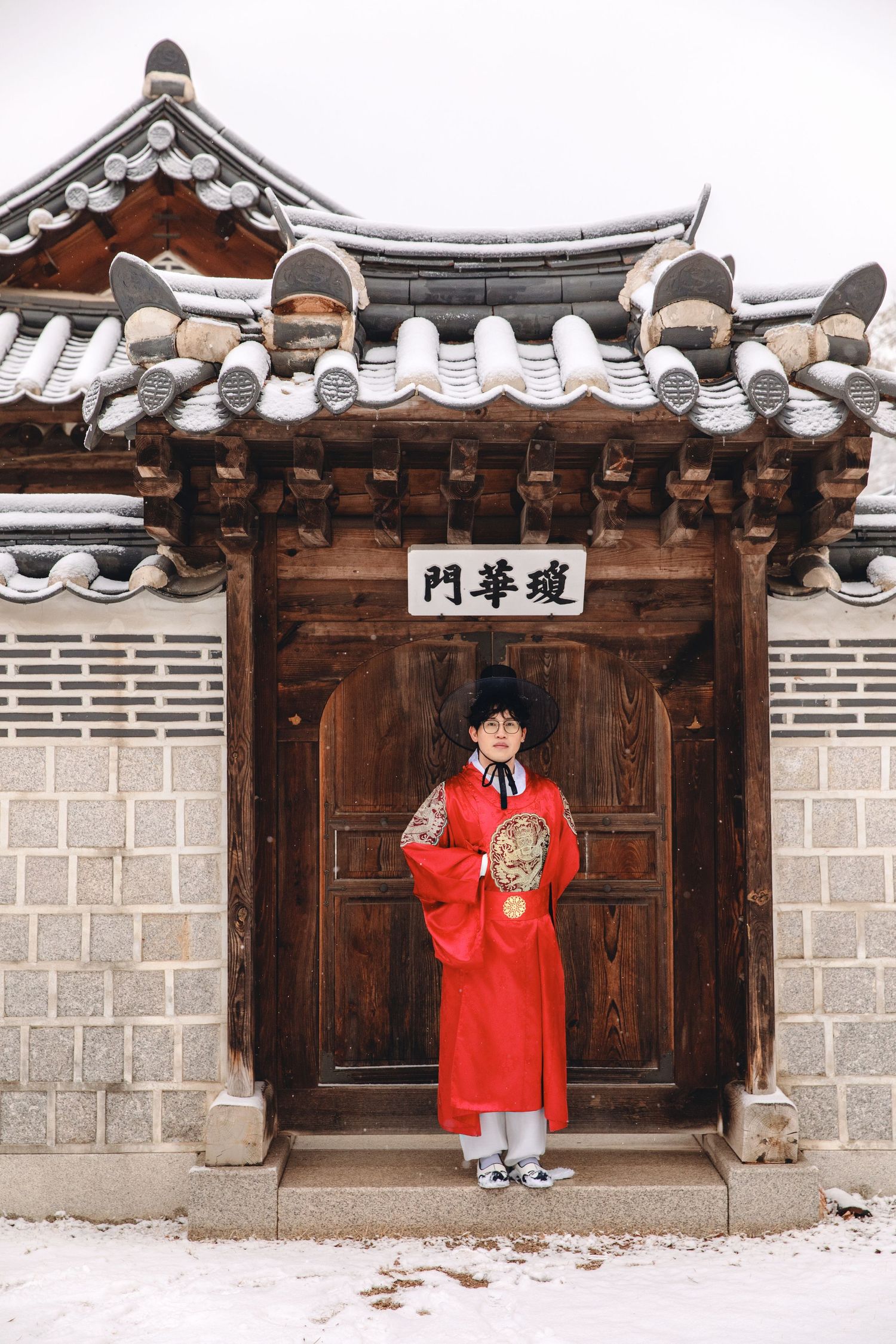 Traditional Korean architecture with curved roof tiles and wooden door frames a figure wearing a bright red hanbok in winter.