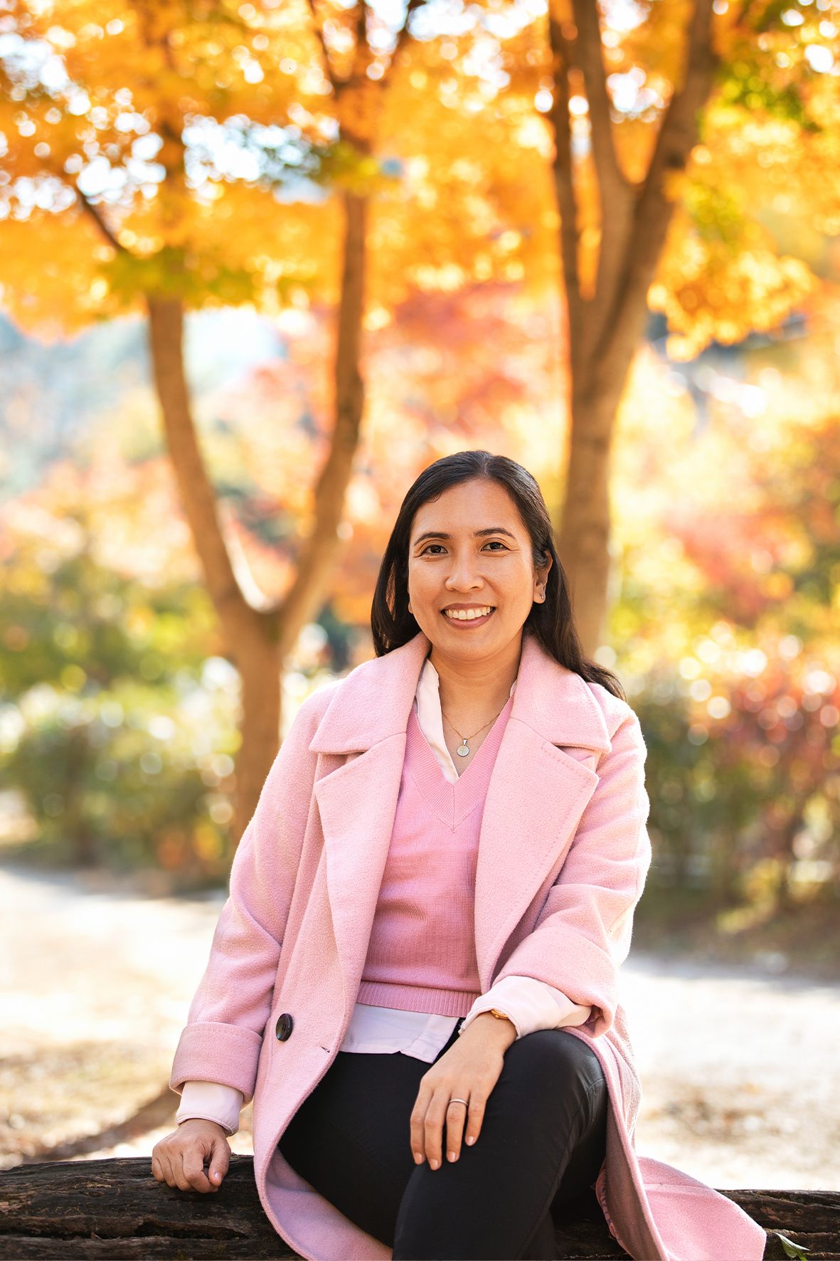 Person in pink coat sits on log against vibrant autumn foliage background in park setting.
