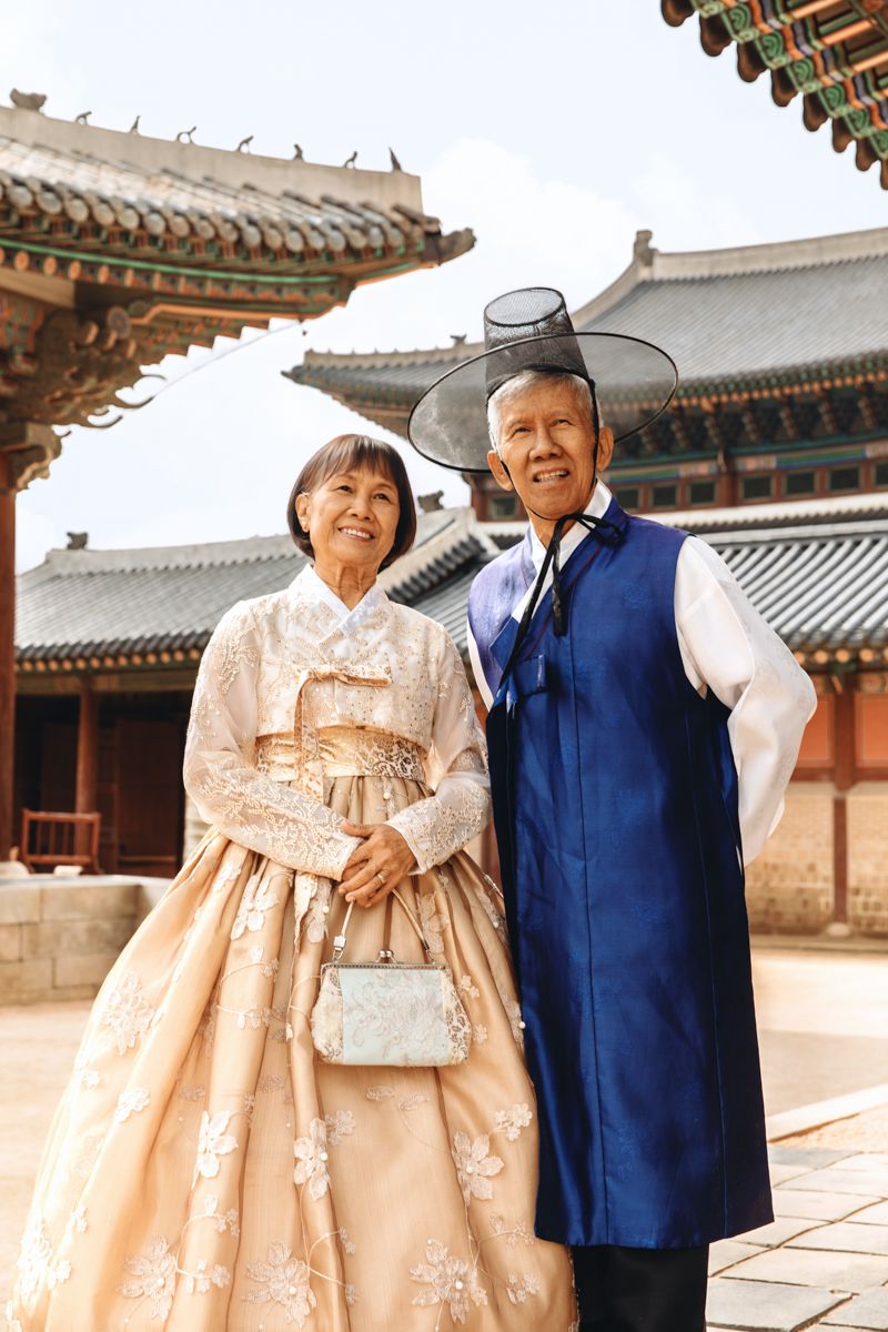 Couple wearing traditional Korean hanbok clothing and a cowboy hat pose at Gyeongbokgung Palace courtyard in Seoul.