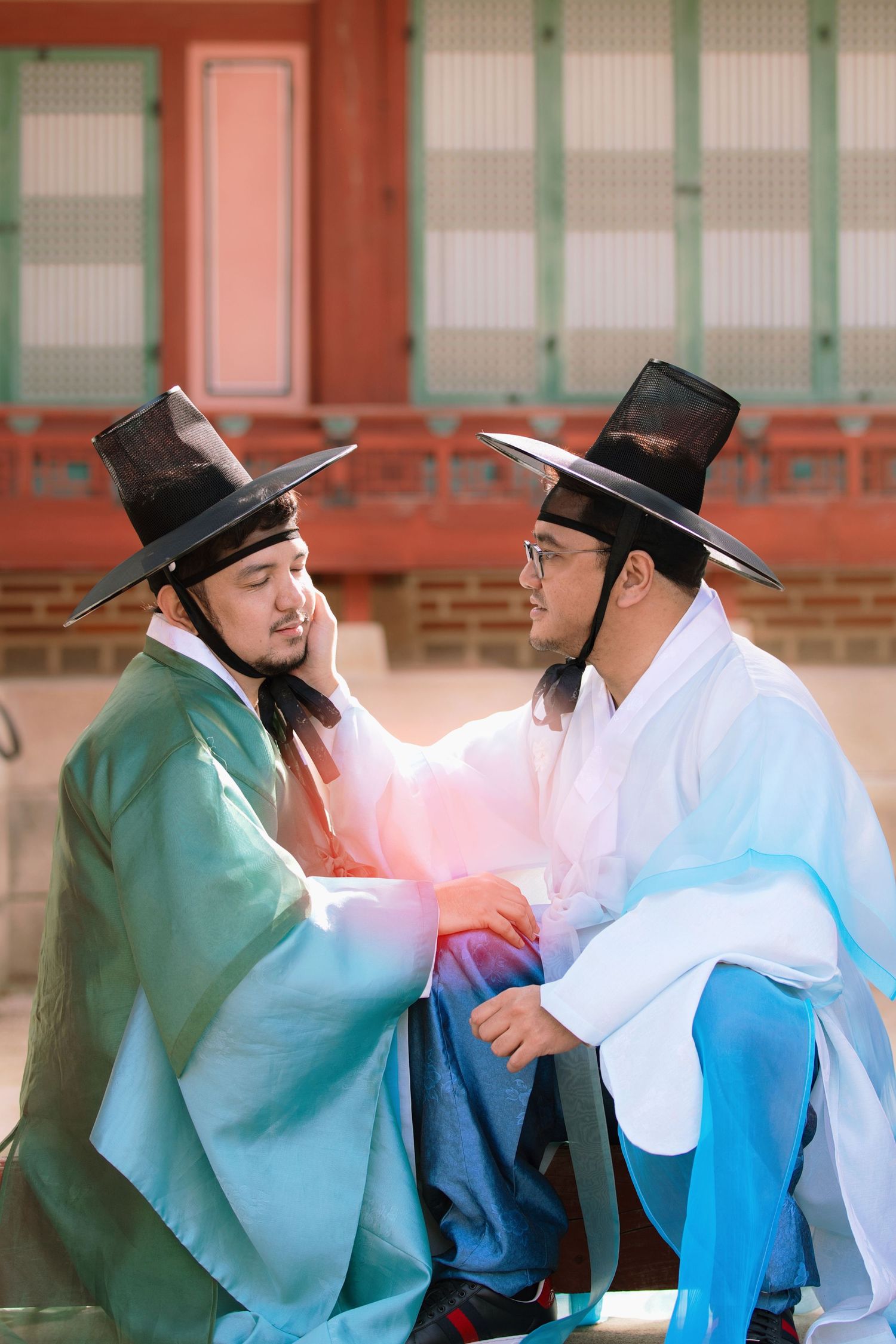 Two lgbt men in traditional Korean hanbok and black hats sitting together on wooden steps at a historic palace Gyeongbokgung Palace in Seoul