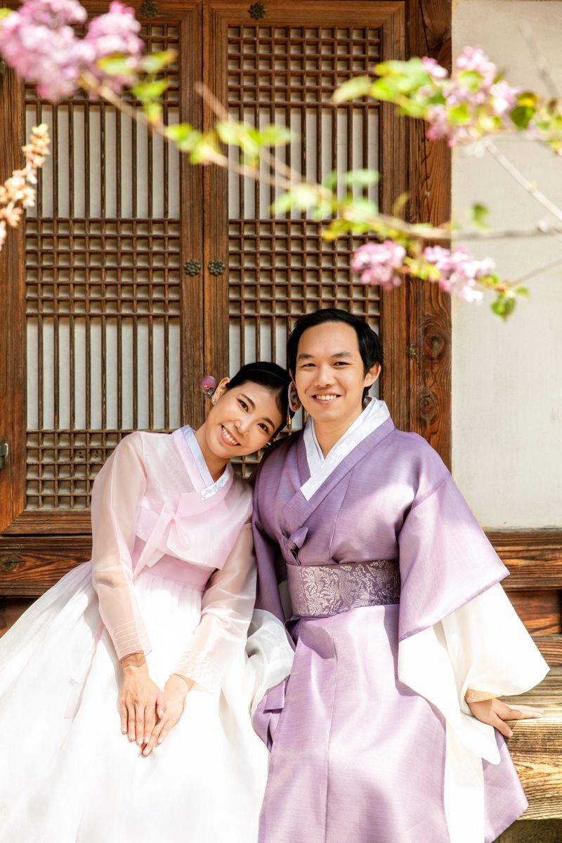 Lovely couple in traditional Korean hanbok sit outside a wooden door with cherry blossoms in spring smiling and sharing love