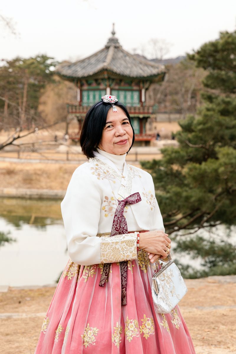 Person wearing traditional Korean hanbok dress standing near a pagoda and lake in a tranquil garden setting in Gyeongbokgung Palace Seoul