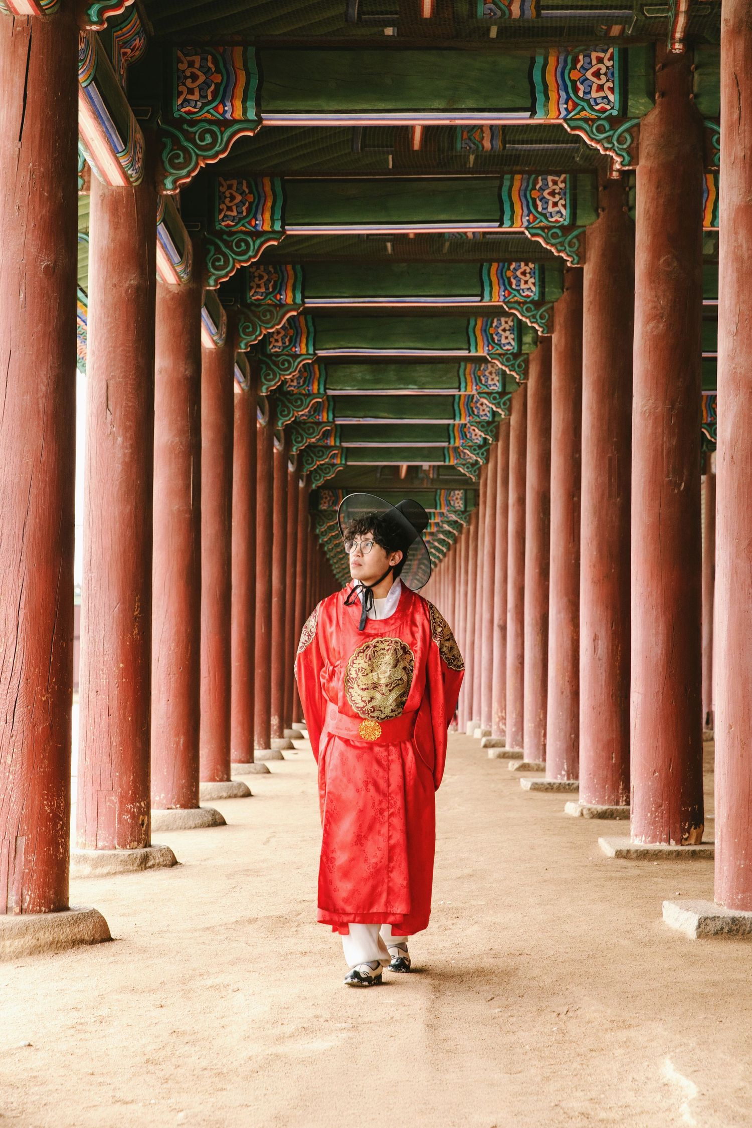 Traditional Korean architecture with red columns and decorative beams creates a covered walkway with someone in red hanbok dress.