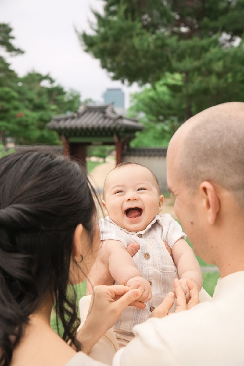 Joyful baby laughs while being held outdoors near a traditional Korean pavilion with trees in the background during the photoshoot