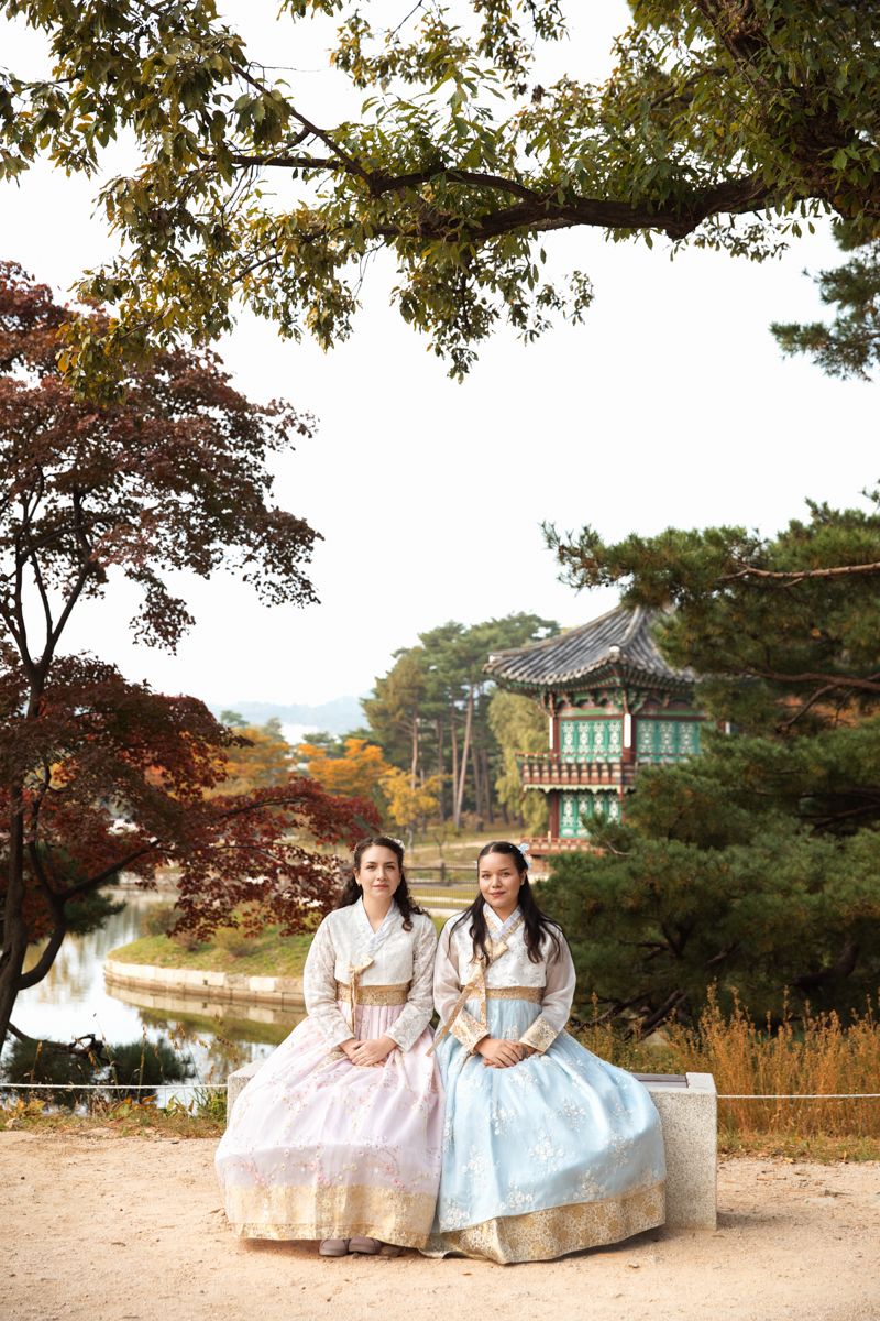 Sisters wearing traditional Korean hanbok sitting against autumn foliage landscape with pagoda and trees at scenic garden setting in Seoul