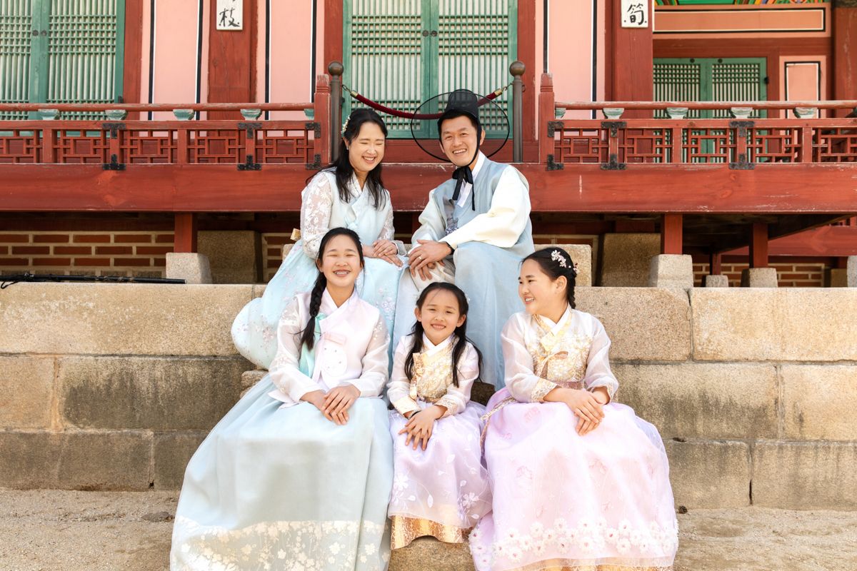 A family wearing traditional Korean pastel hanbok attire pose together on stone steps in front of historic Gyeongbokgung Palace in Seoul