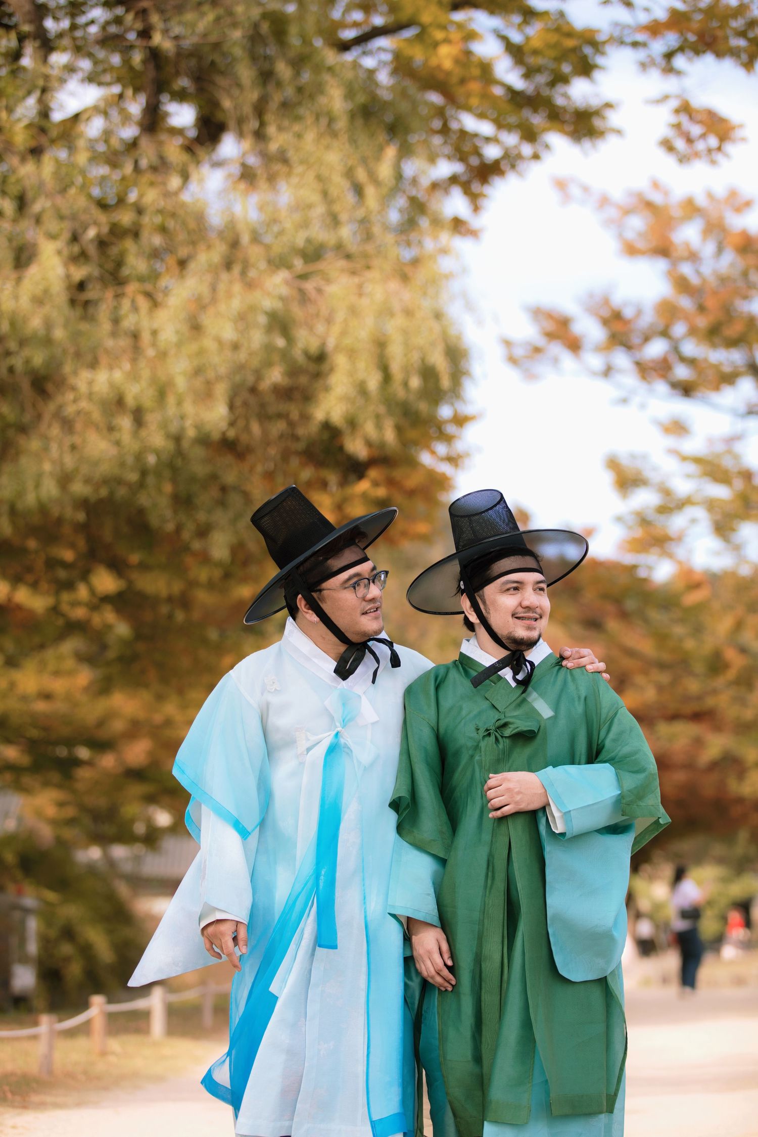 Two people wearing traditional Korean hanbok outfits and black hats walk together on an autumn path surrounded by trees in Gyeongbokgung