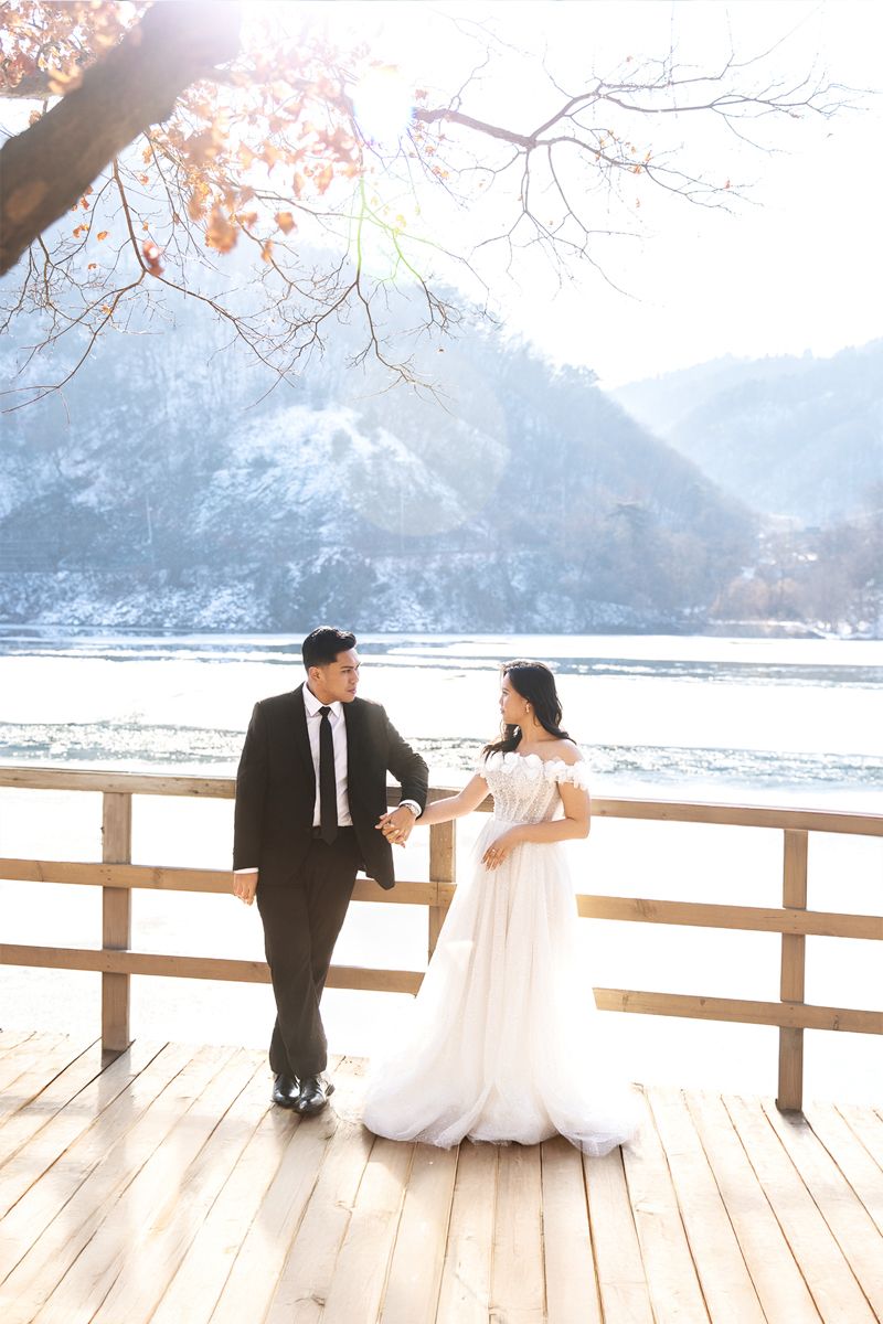 A romantic winter wedding couple sits on a wooden deck overlooking a snowy mountain lake during their photo session.