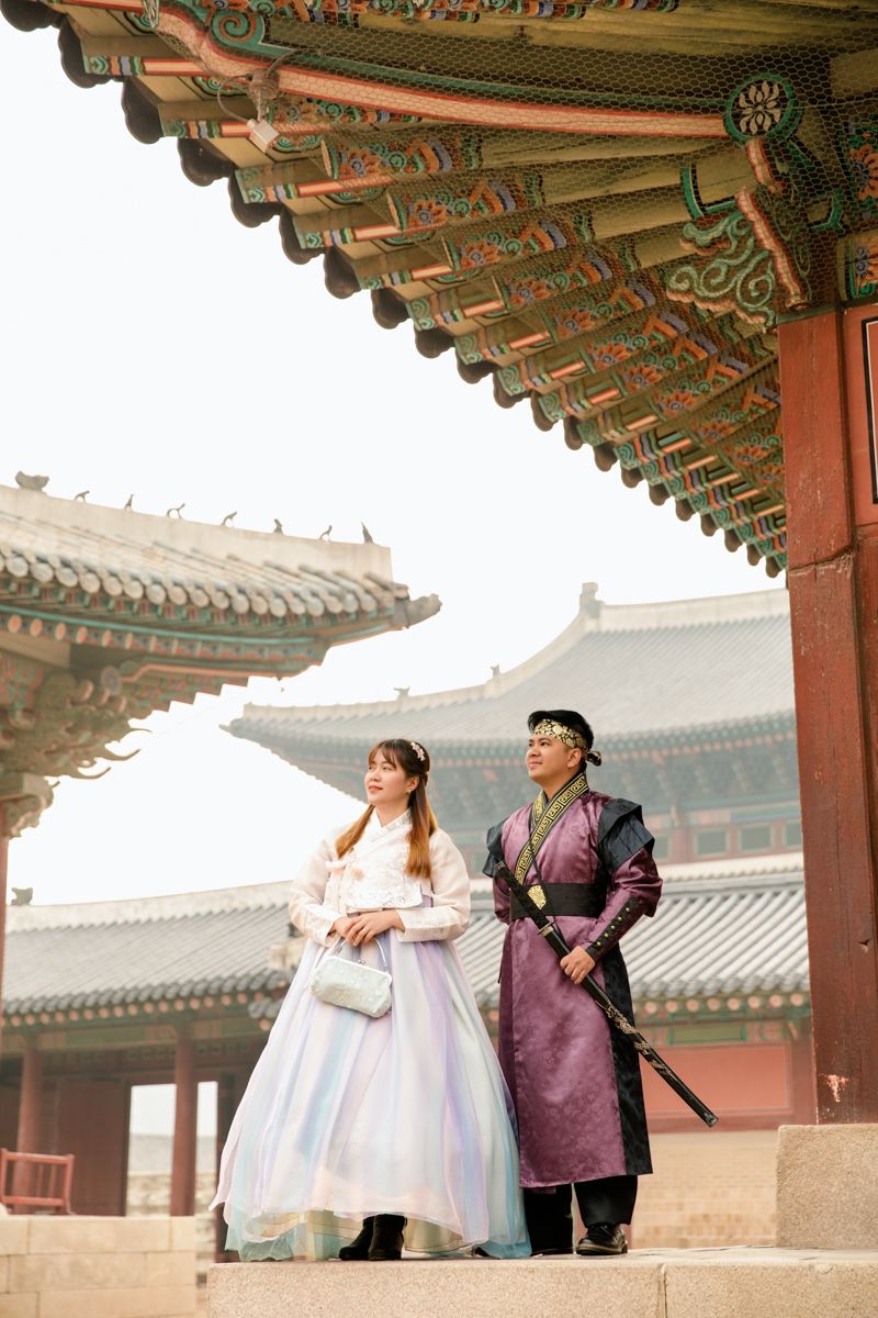 Romantic couple wearing traditional Korean hanbok attire stand under ornate wooden Gyeongbokgung Palace in Seoul. Smiling