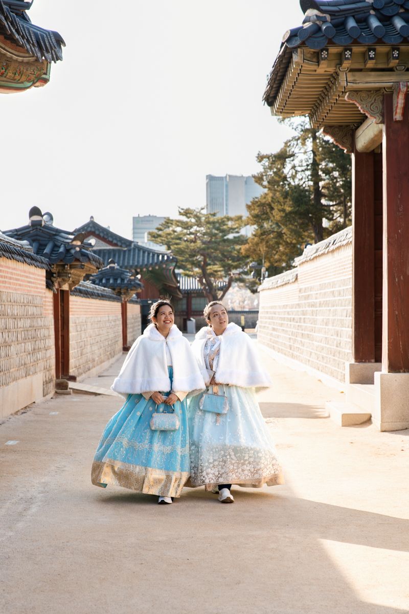Two girls wearing traditional blue Korean hanbok walking through a historic Gyeongbokgung palace courtyard with traditional architecture.