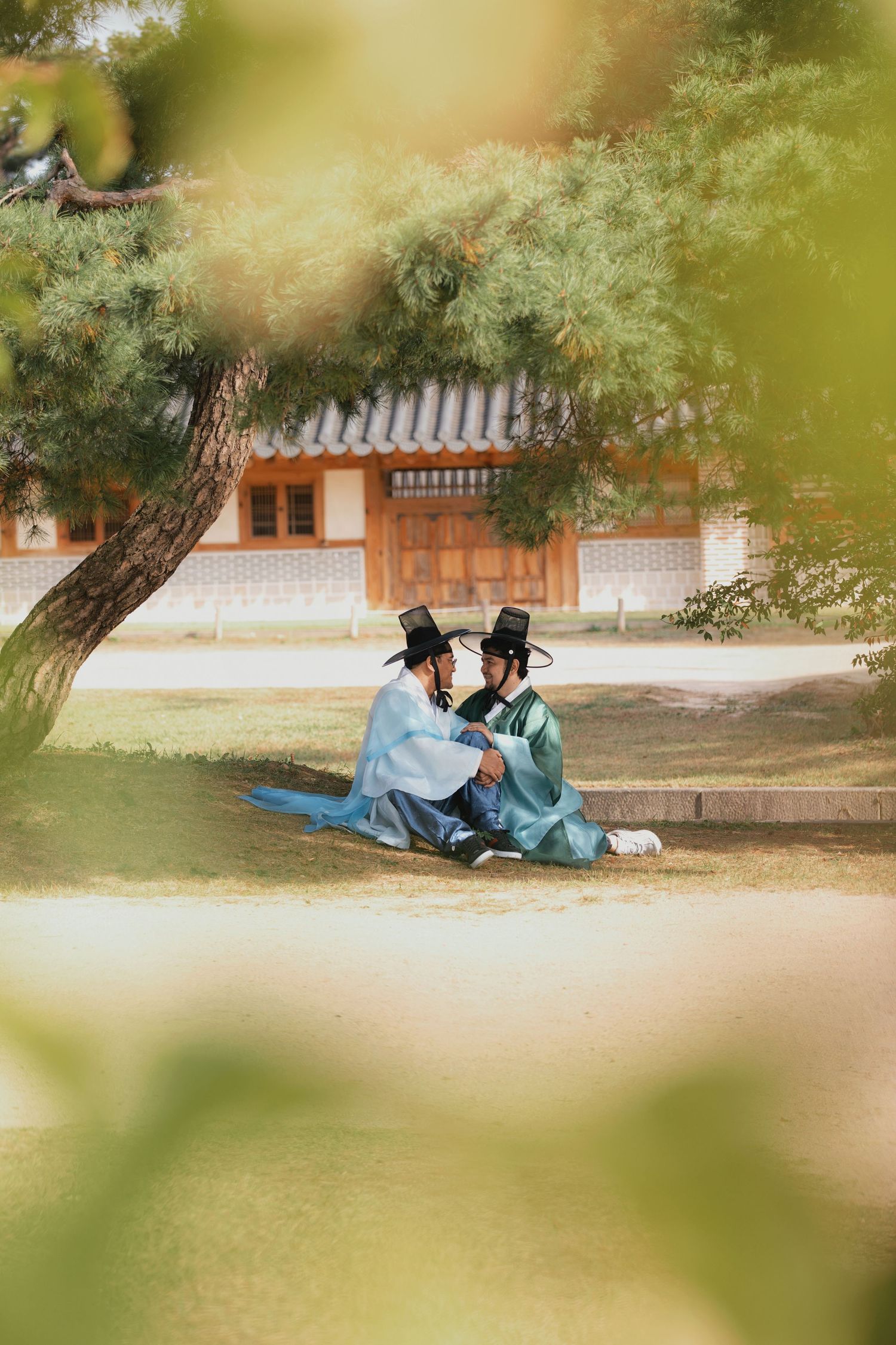 Two people sit together on grass near historic buildings under tree shade in a tranquil outdoor setting.