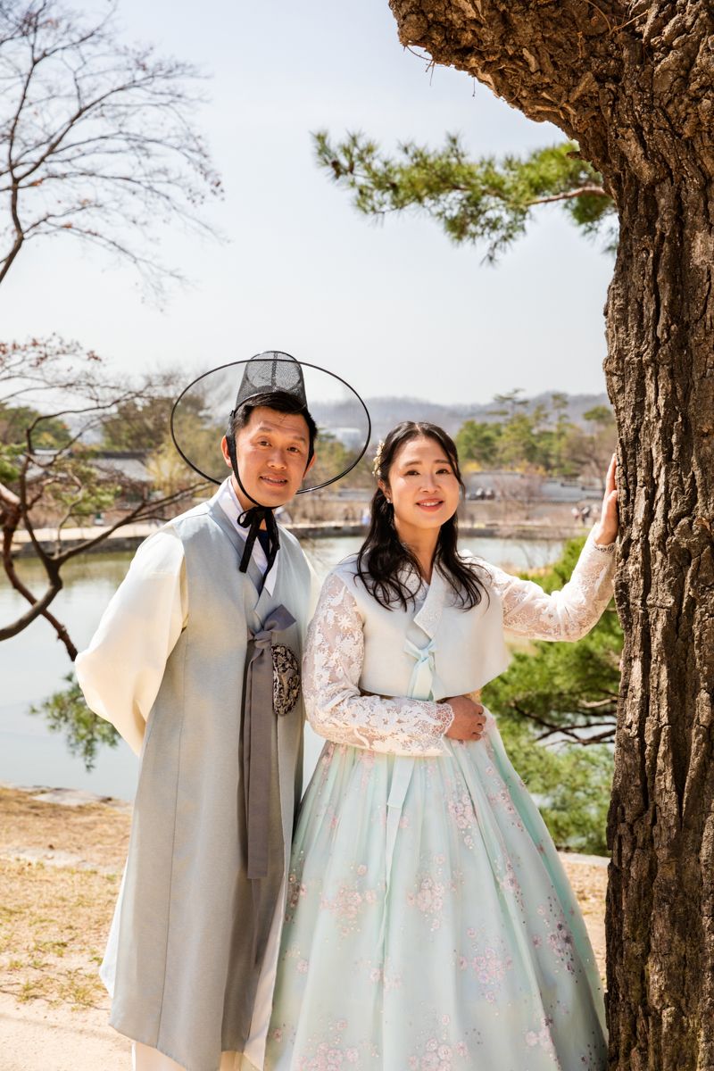 Two people in traditional Korean hanbok attire pose together by a tree during springtime.