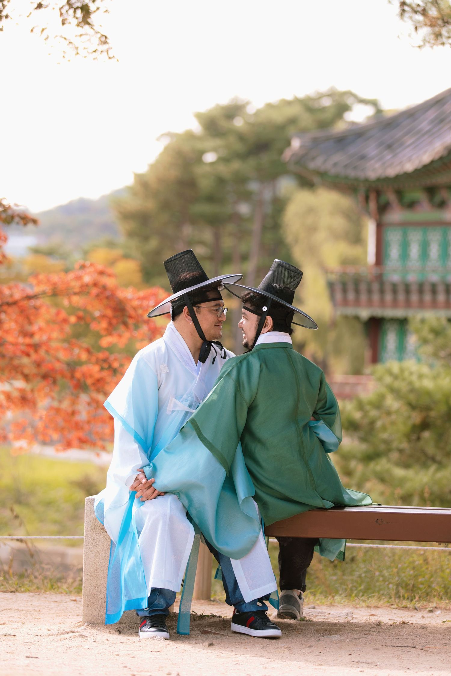 Two people in traditional Korean hanbok attire and black hats sit together on a bench during autumn with red foliage in South Korea