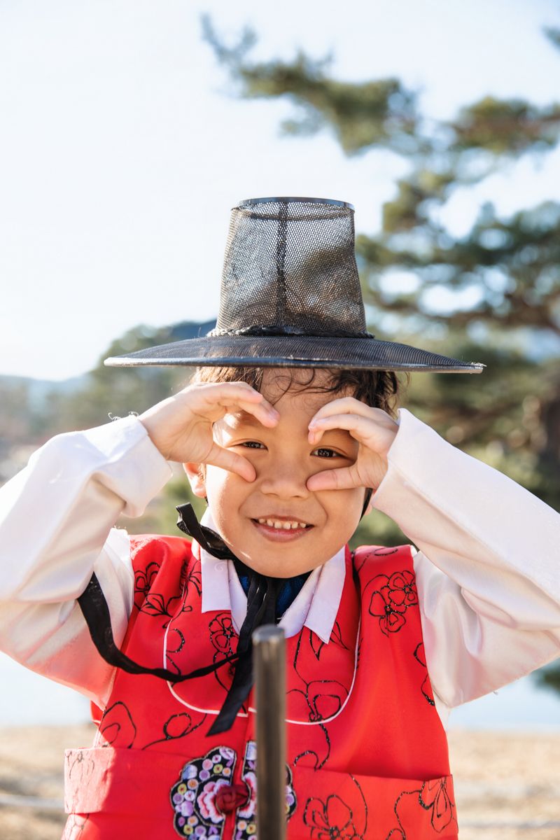 Boy wearing red hanbok with red vest and tall black hat makes playful gesture with hands in sunlit outdoor setting in Gyeongbokgung Palace