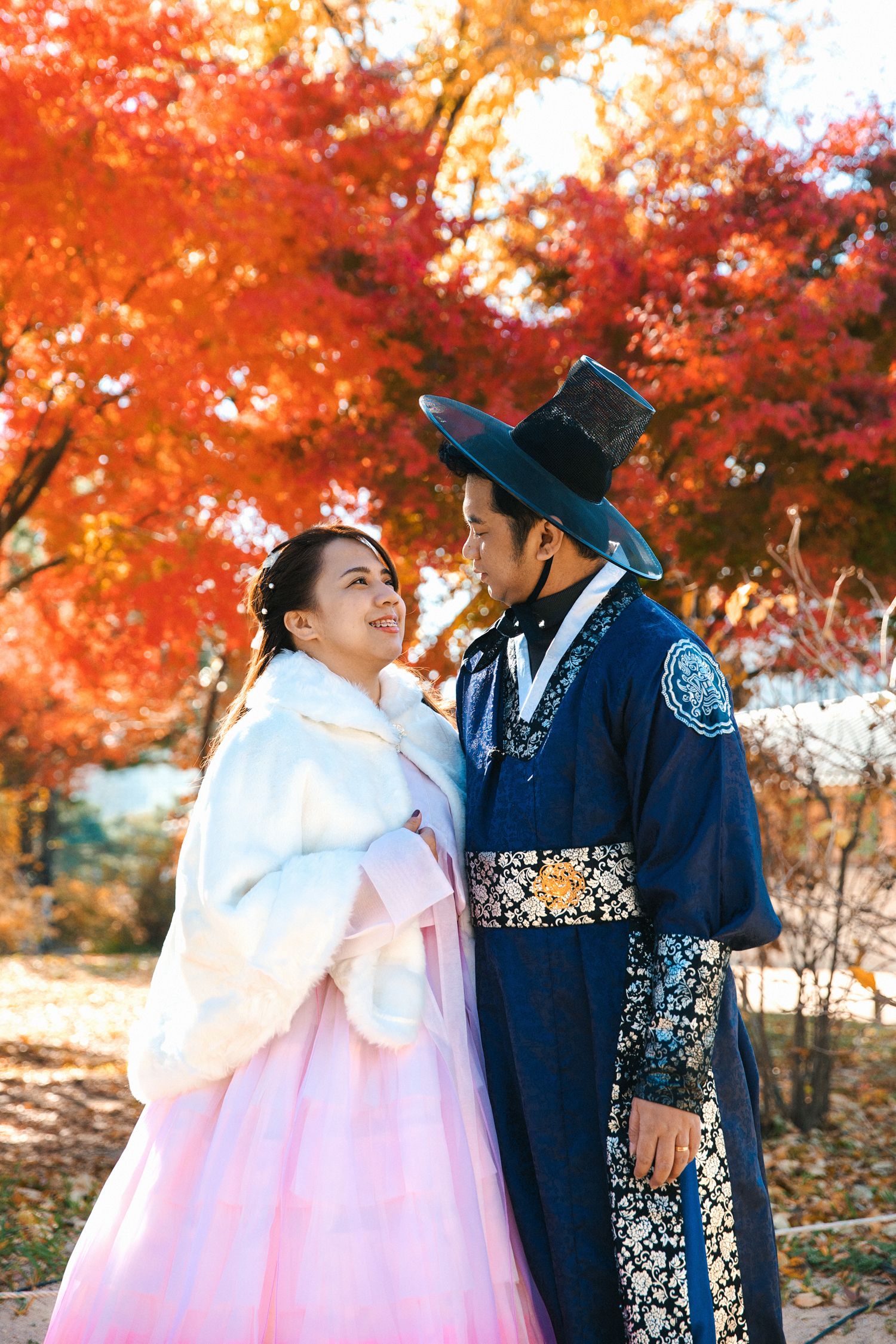 Couple in traditional Korean hanbok poses beneath vibrant red autumn maple trees.
