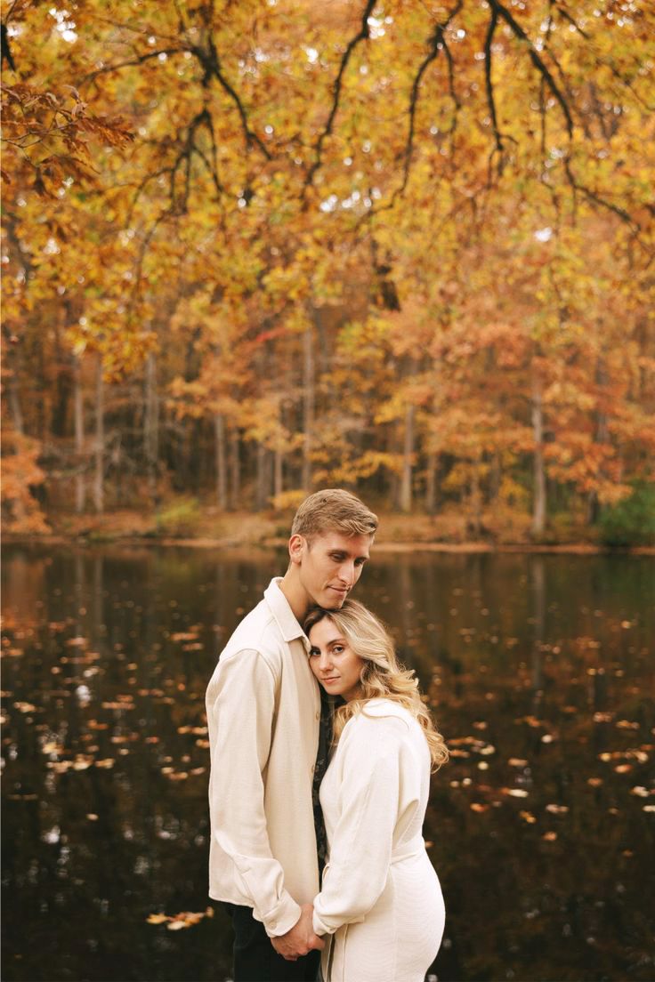 Two figures in white outfits embrace against a backdrop of autumn foliage and a tranquil lake with fallen leaves.