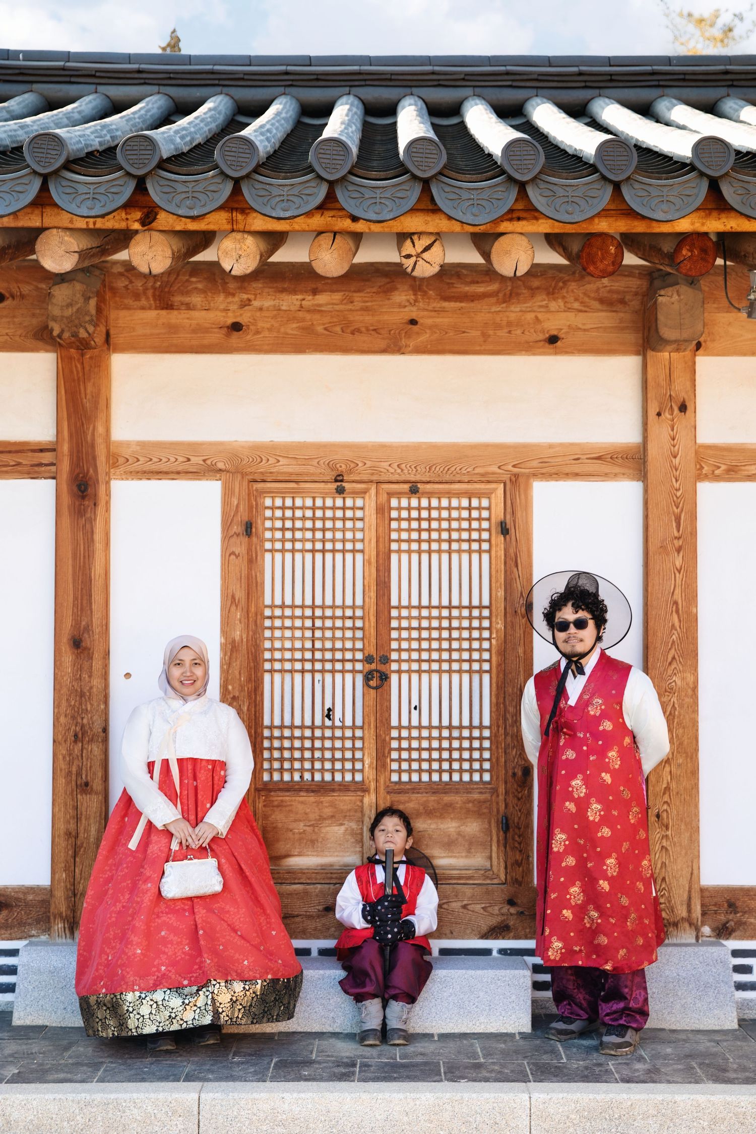 Traditional Asian family wearing vibrant red hanbok outfits pose in front of ornate wooden doorway at historic building.
