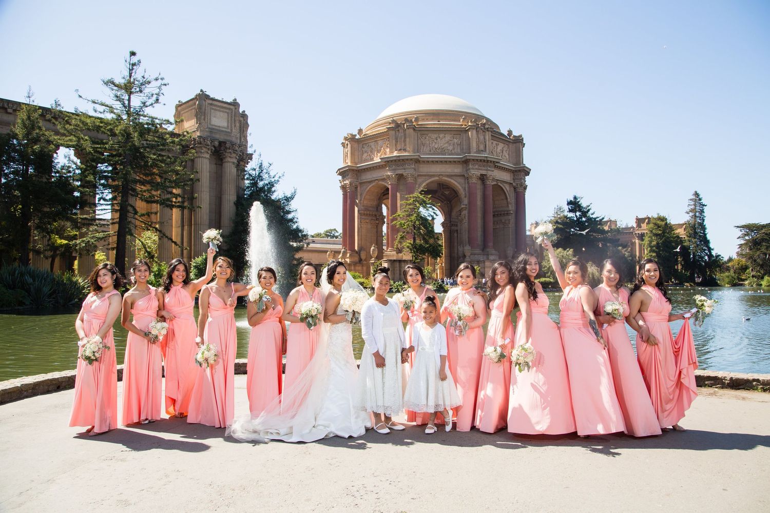 Bridal party wearing pink dresses poses at the Palace of Fine Arts with fountains and architecture.