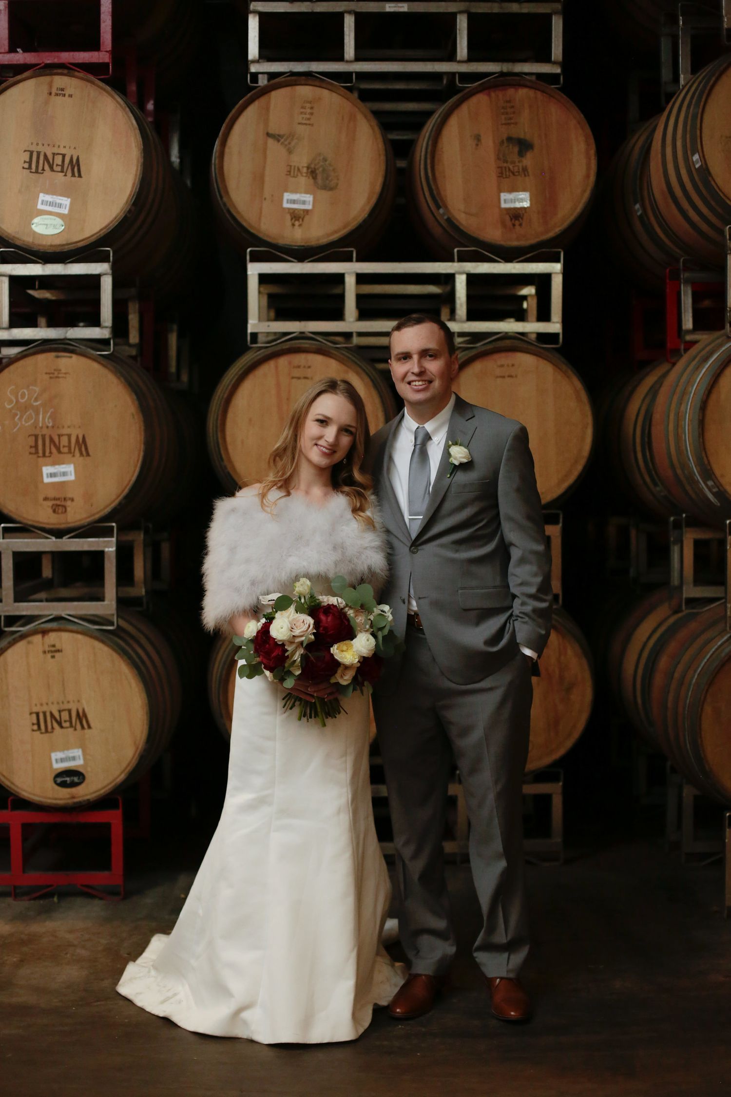Wedding portrait in front of stacked wine barrels in rustic winery setting.