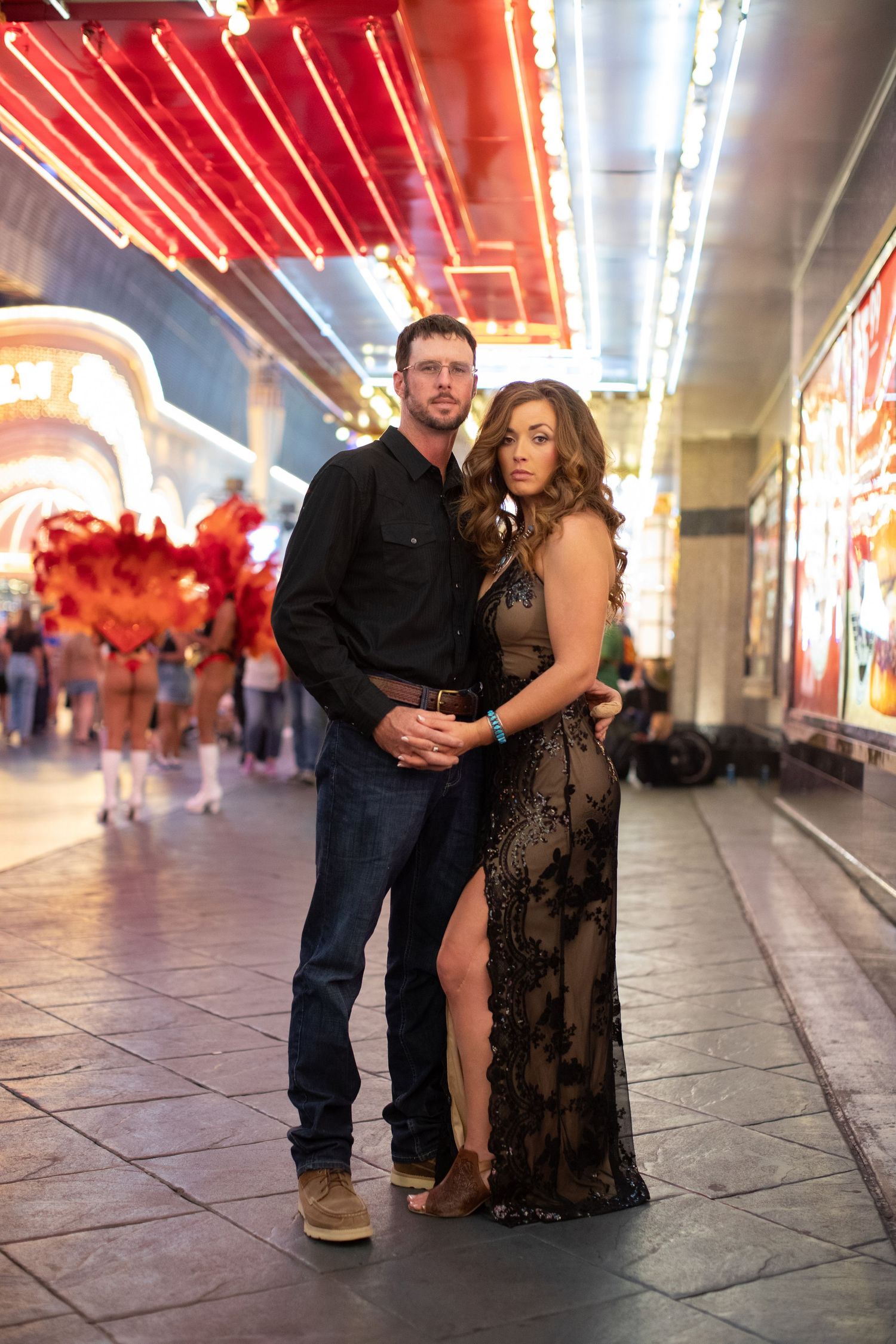 Couple poses intimately under neon lights in an elegant black dress and casual attire on city street.