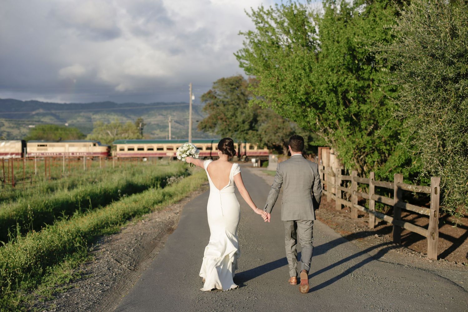 A couple walks hand in hand down a country road with mountains in the background.