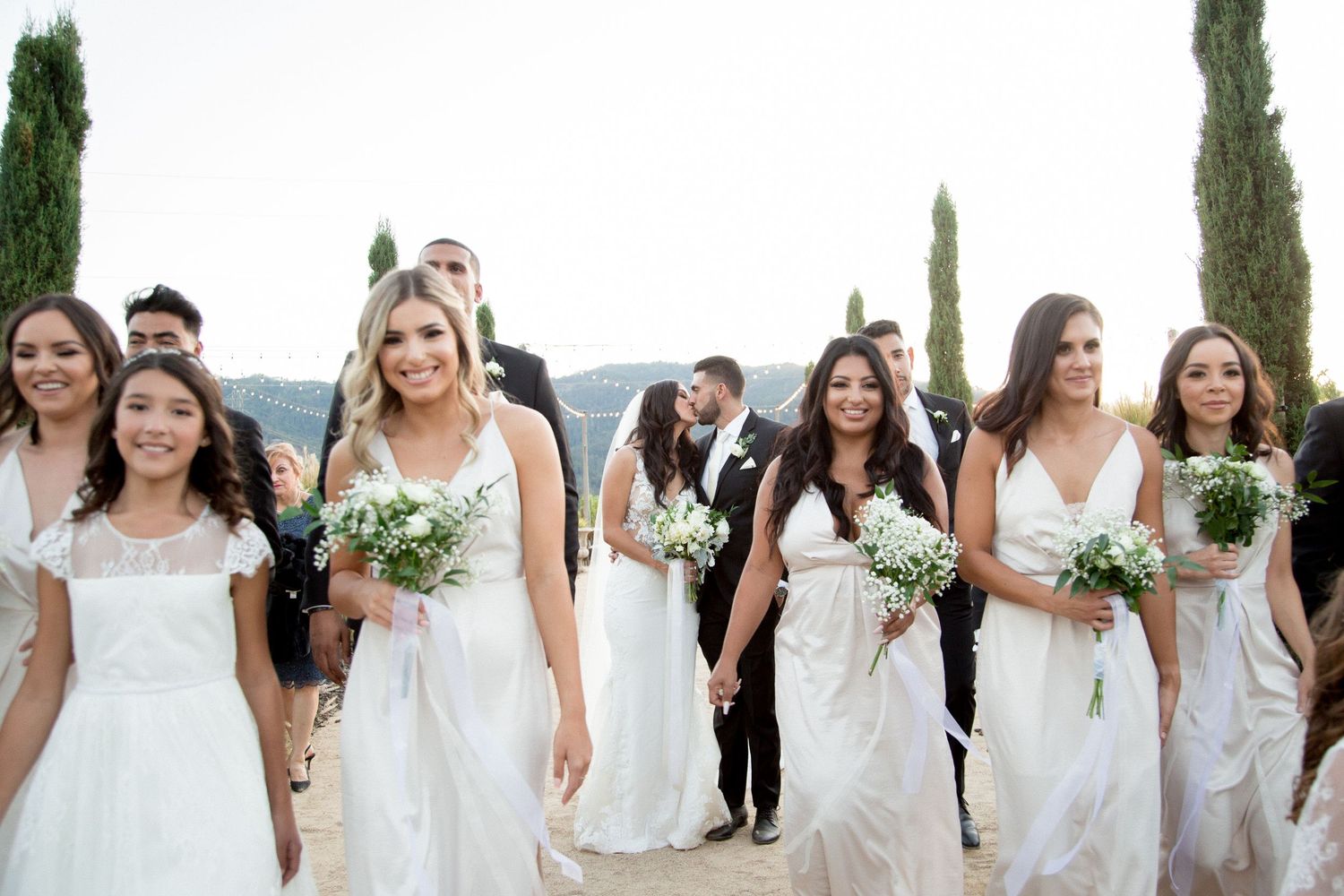 Bridal party in flowing white dresses gather for group photo outdoors.