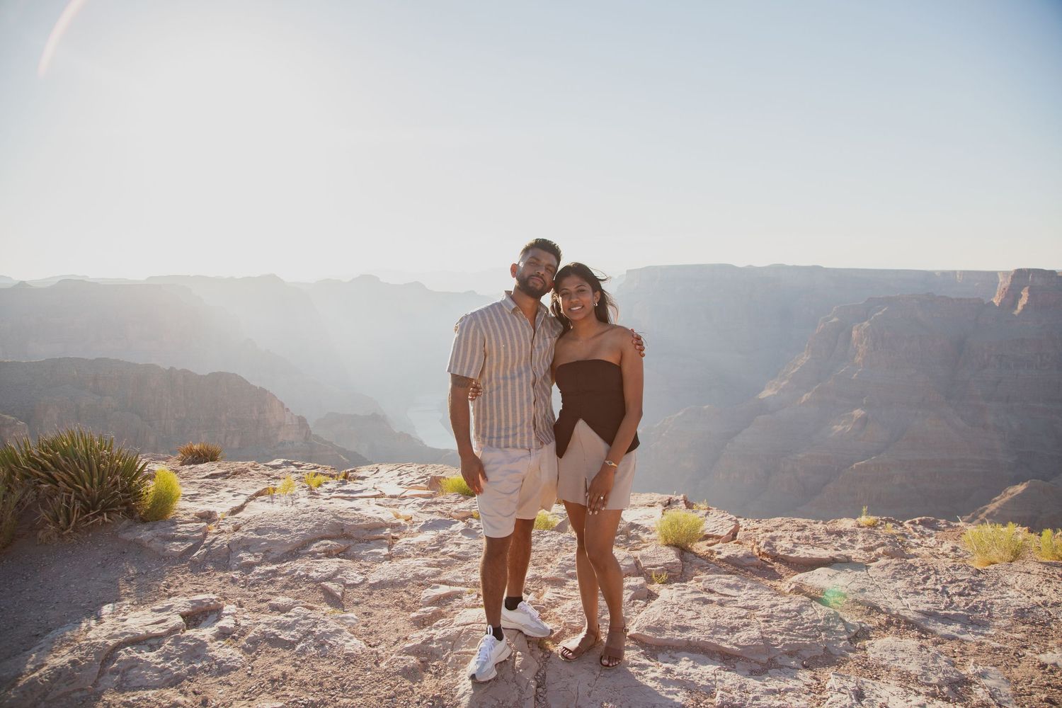 A couple stands together at the edge of the Grand Canyon during sunset with mountains in the background.