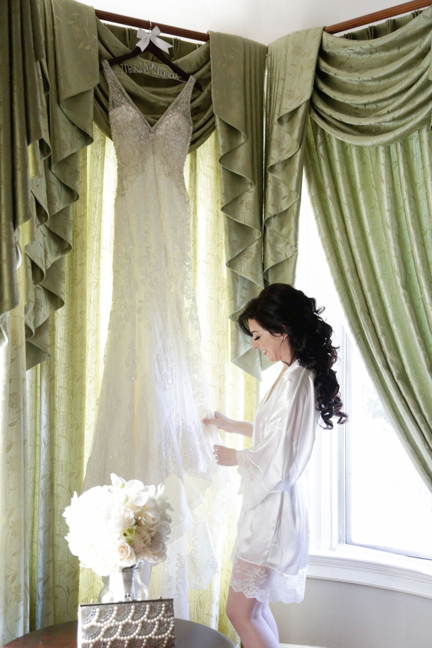 Bride in white robe getting ready by elegant draped windows.