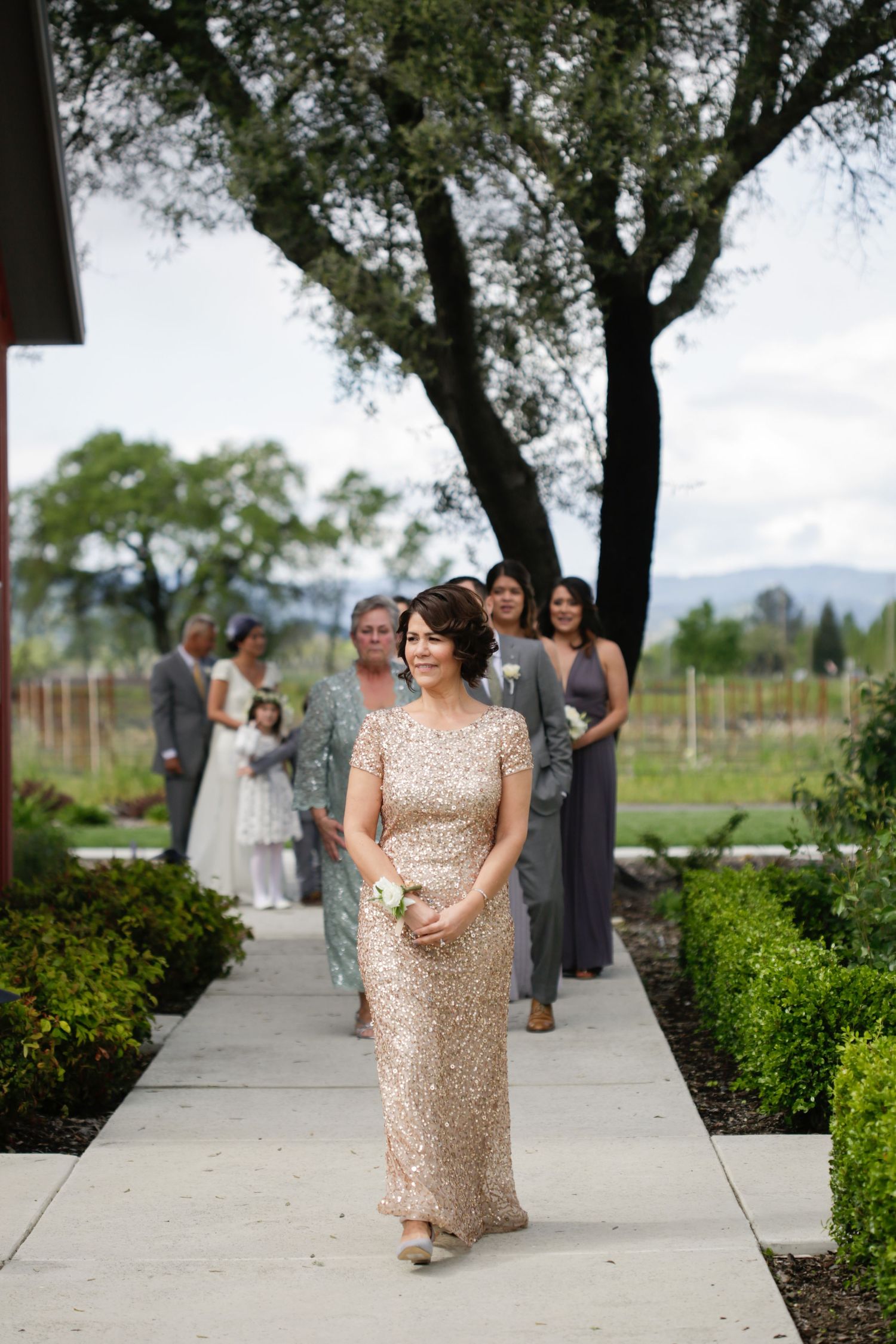 Wedding attendant in a sparkly gold dress walks down an outdoor pathway lined with trees.