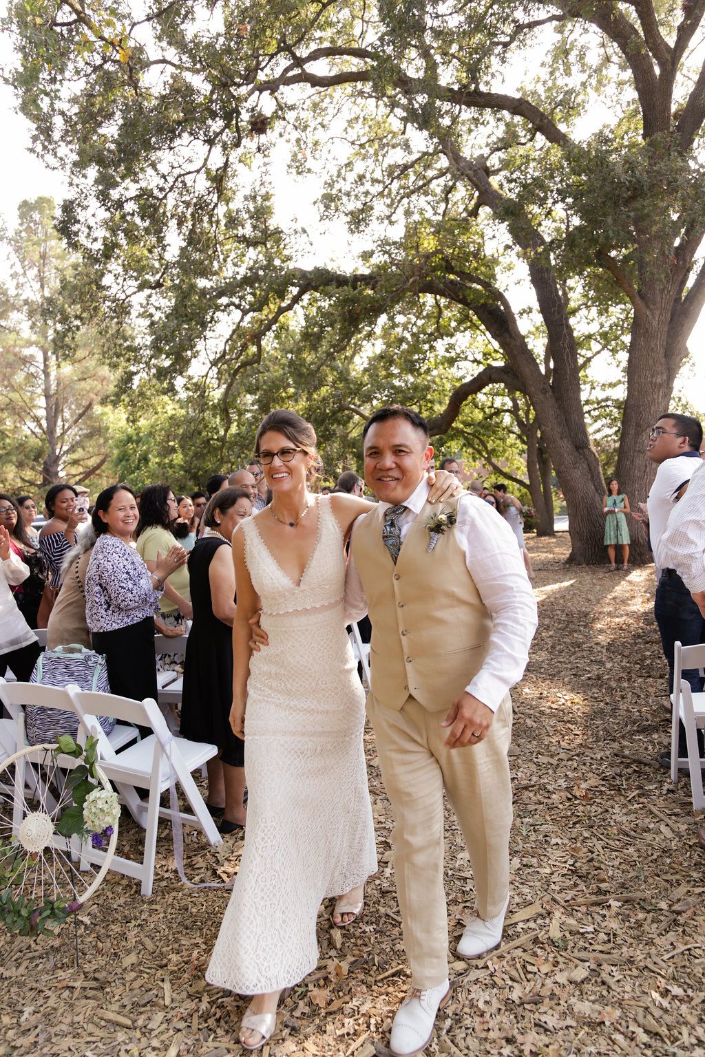 Wedding party celebrating outdoors under large oak trees with white chairs.