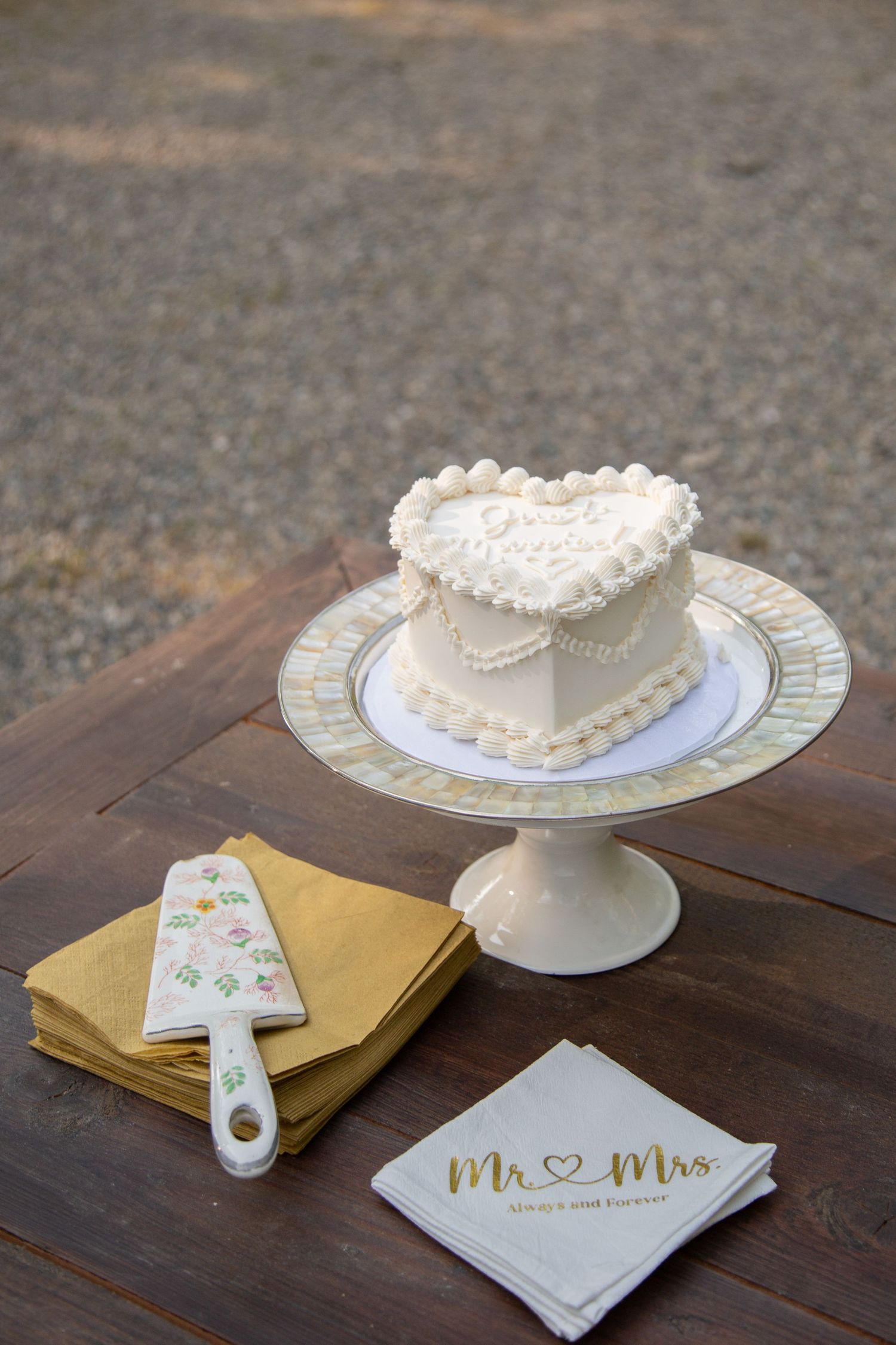 Small heart-shaped wedding cake displayed on white pedestal with decorative plates and napkins.