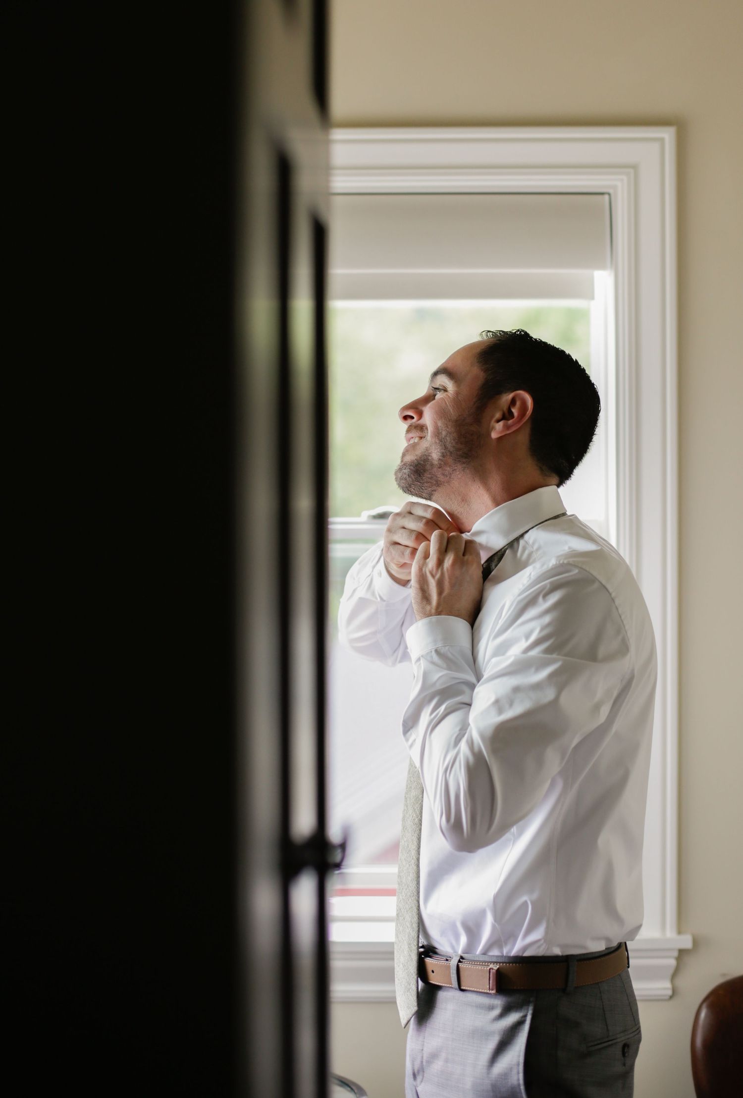 A person in white dress shirt adjusts their tie while standing by a window.