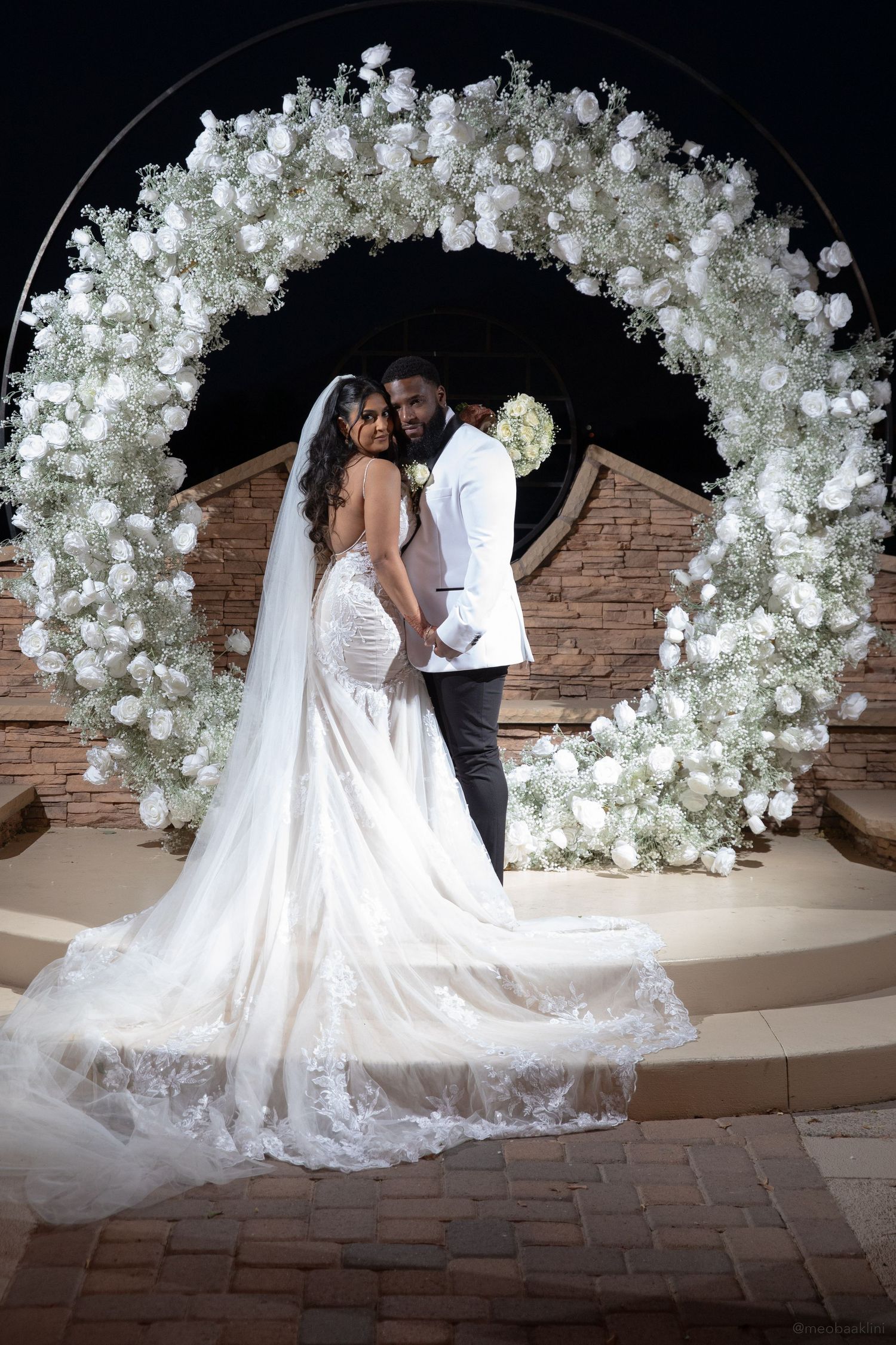 Newlyweds pose beneath an ornate arch covered in white flowers and greenery.
