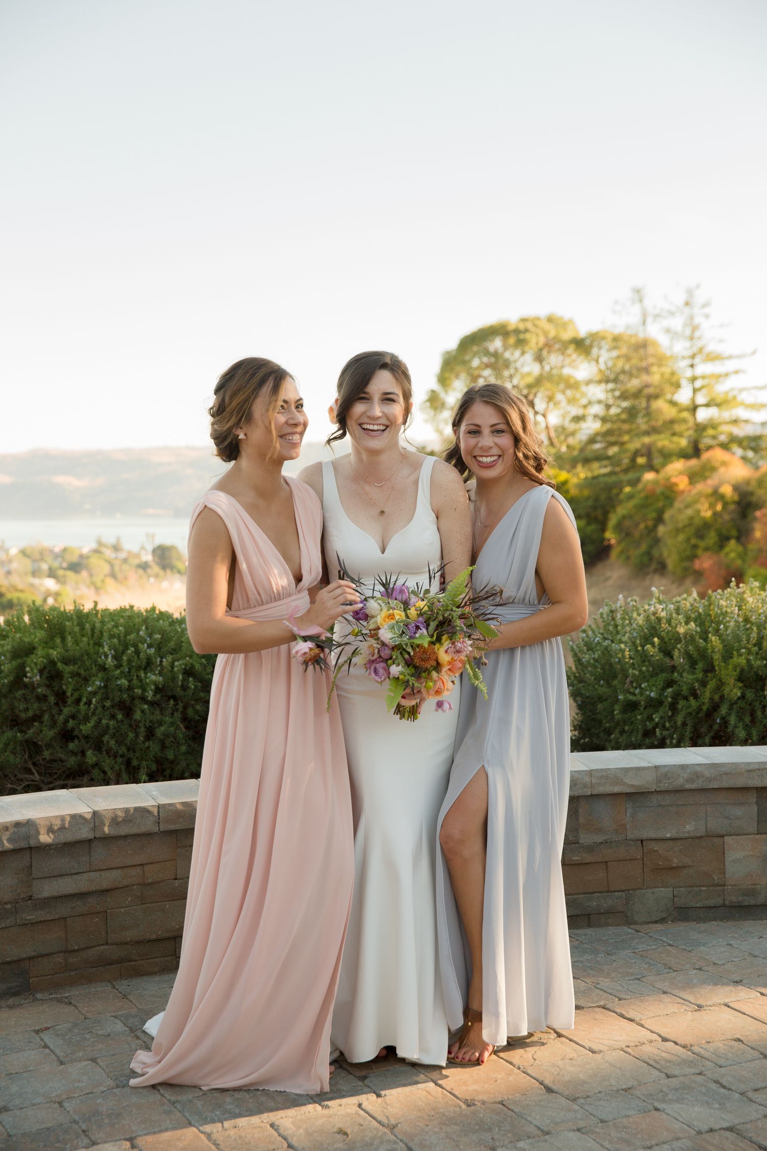 Three women in elegant dresses pose outdoors at sunset holding bouquets.